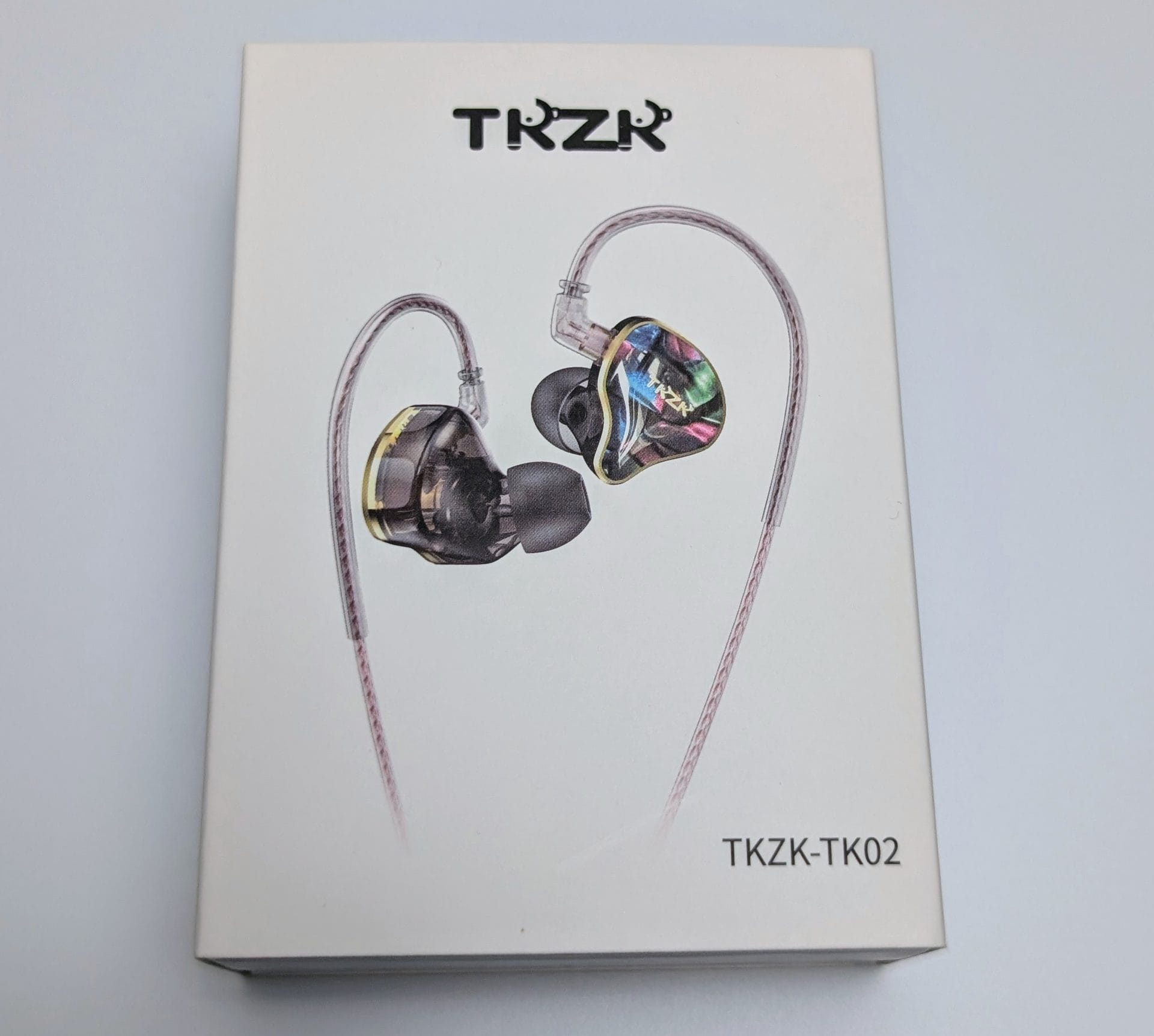 TKZK TK02 IEMs The image shows the packaging of the TKZK-TK02 in-ear monitors (IEMs). The box features a white background with a central illustration of the IEMs, highlighting their transparent shells and colorful, reflective faceplates. The brand name "TKZK" is displayed prominently at the top, and the model name "TKZK-TK02" is written at the bottom.