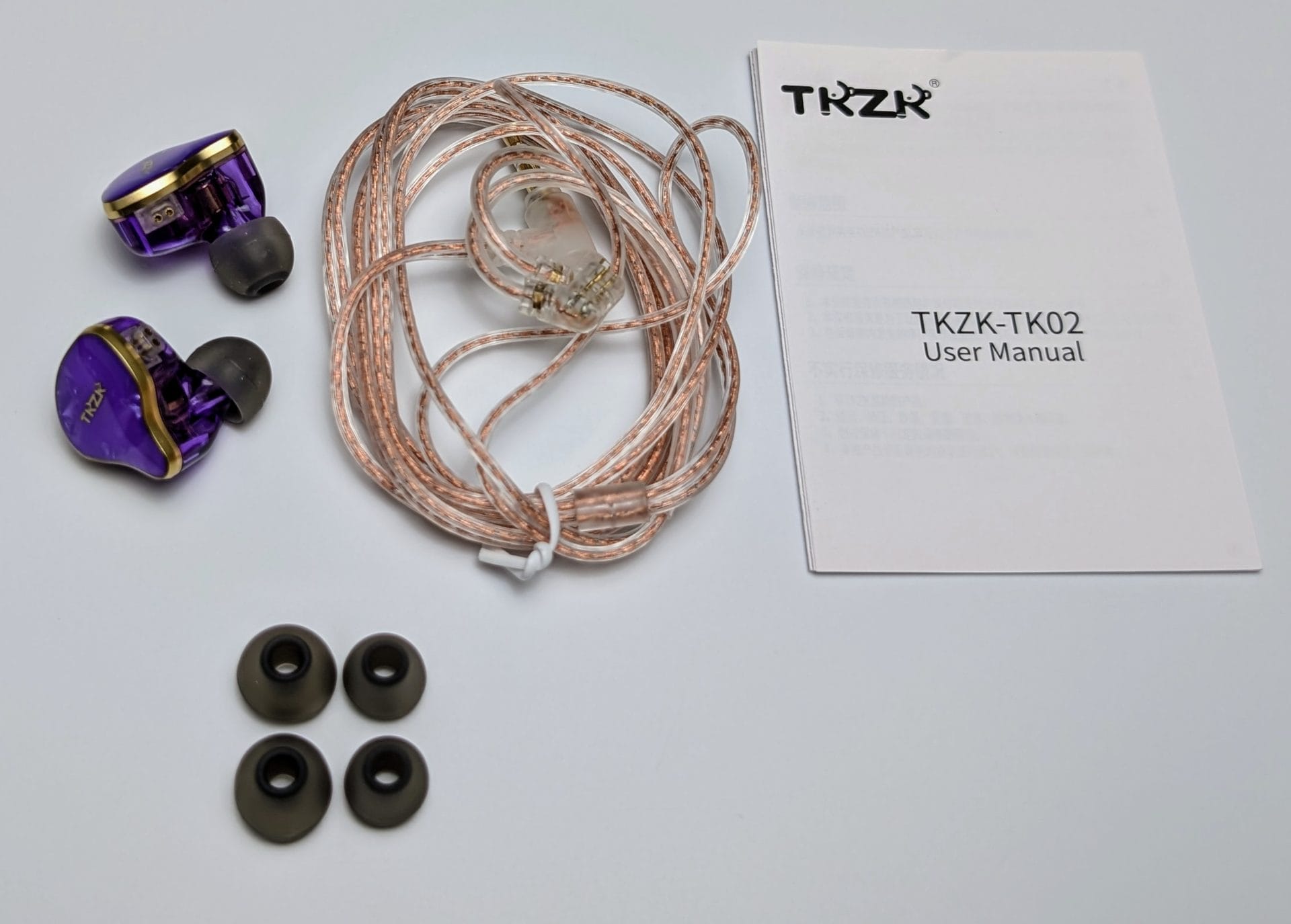 TKZK TK02 IEMs The image displays the contents of the TKZK-TK02 IEM package. Included are:1. A pair of purple in-ear monitors with gold accents. 2. A detachable transparent braided cable with 2-pin connectors. 3. Three pairs of silicone ear tips in different sizes (small, medium, and large). 4. A user manual titled "TKZK-TK02 User Manual." The items are neatly arranged against a white background.