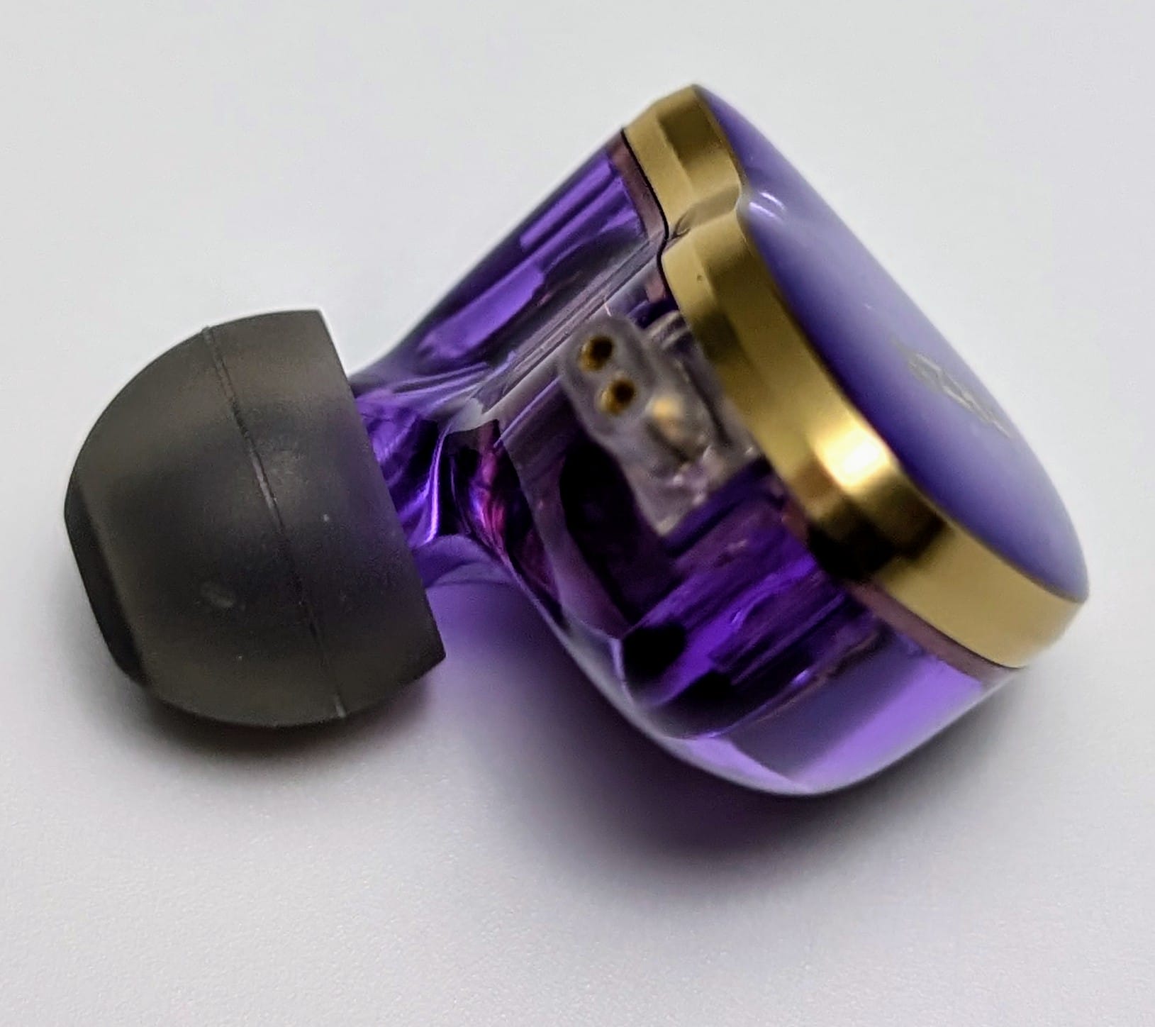 TKZK TK02 IEMs A close-up of a sleek, modern earbud in a vibrant purple colour with gold accents. The earbud has a glossy finish and features a black ear tip. The design appears sophisticated and high-tech, with a partial view of the internal components visible through the translucent casing.