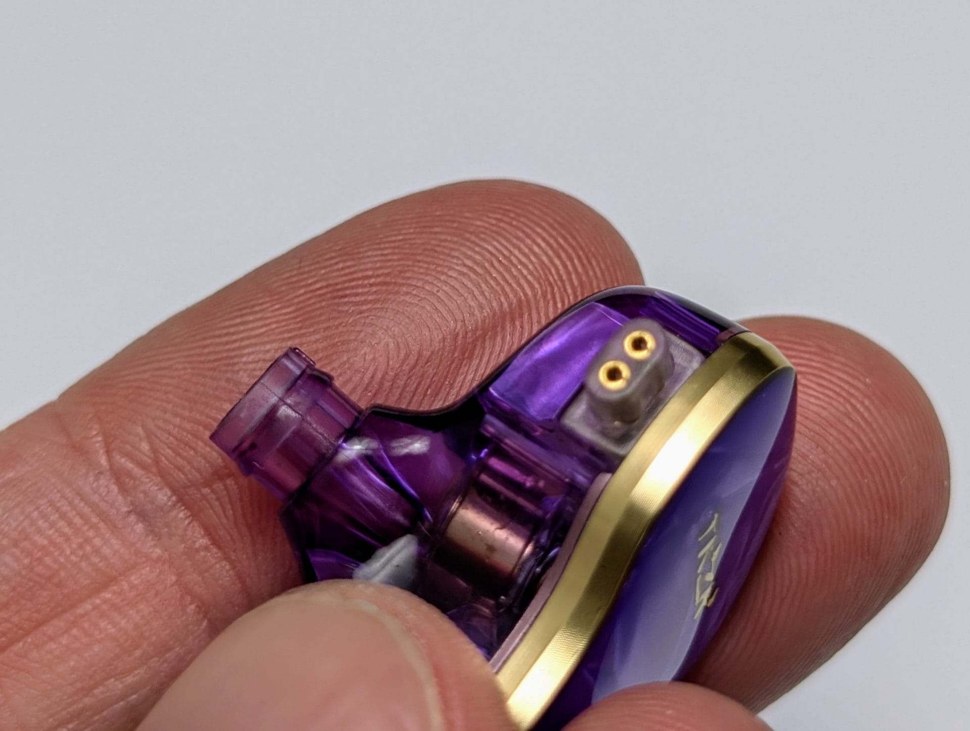 TKZK TK02 IEMs Close-up of a hand holding a single purple in-ear monitor (IEM) with gold accents. The IEM has a distinct, shiny, metallic purple finish with the brand name 'TKZK' partially visible. Two gold-coloured connector pins are visible, indicating the earbud's connection point.
