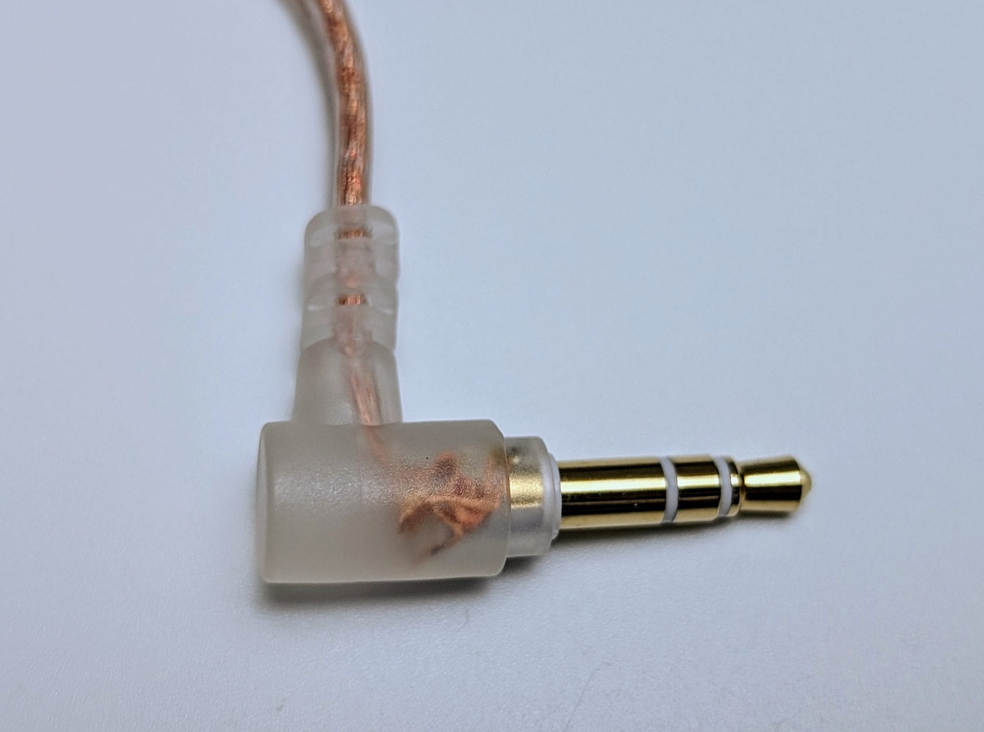 TKZK TK02 IEMs Close-up of a 3.5mm audio jack connector with a right-angle design. The connector features a gold-plated tip with two rings (indicating a TRS connection), and the housing is made of transparent plastic, revealing the internal copper wiring. The cable attached to the connector is braided and enclosed in a translucent sleeve. The background is plain white.