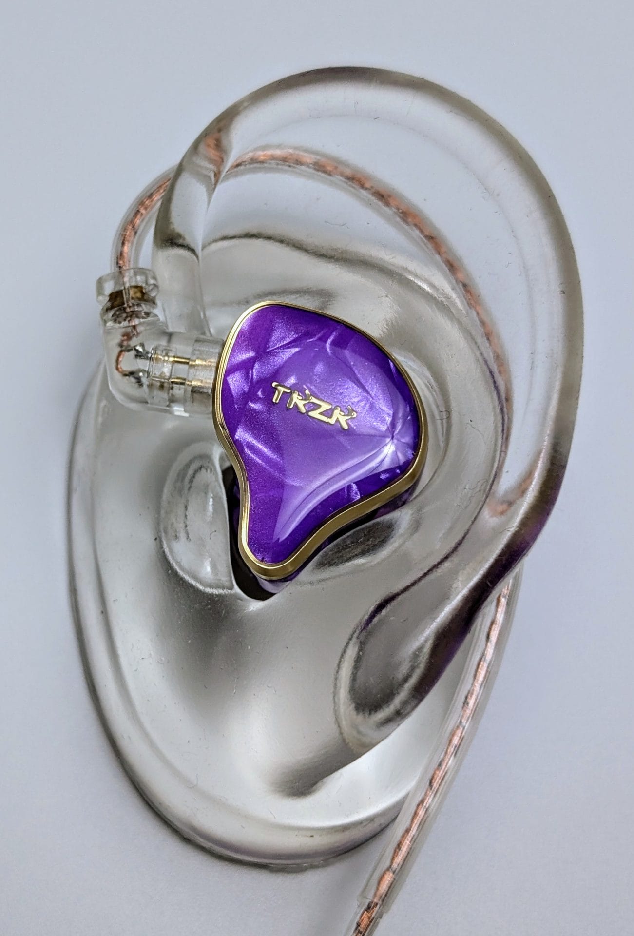 TKZK TK02 IEMs A close-up of an earbud with a shiny purple casing, featuring the brand name 'TKZK' in gold lettering. The earbud is set against a translucent, detailed model of a human ear, highlighting its fit. The earbud's cable is also visible, wrapped around the ear model.