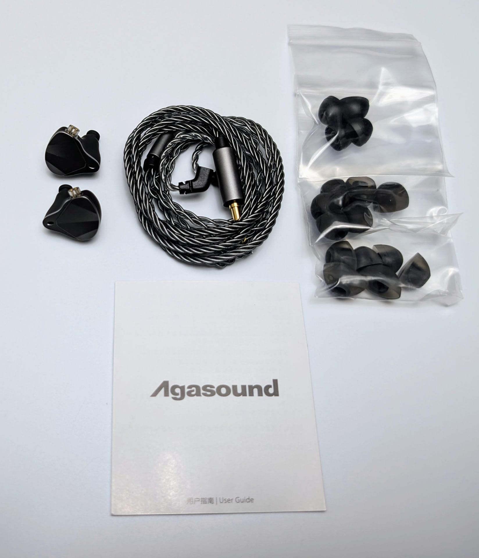 Agasound AgaOne IEMs Image of the contents of the Agasound AgaOne IEMs package. It includes two black in-ear monitors, a coiled black and silver braided cable with a 3.5mm jack, a set of black ear tips in various sizes inside clear plastic bags, and a white user guide with the 'Agasound' logo printed on it.