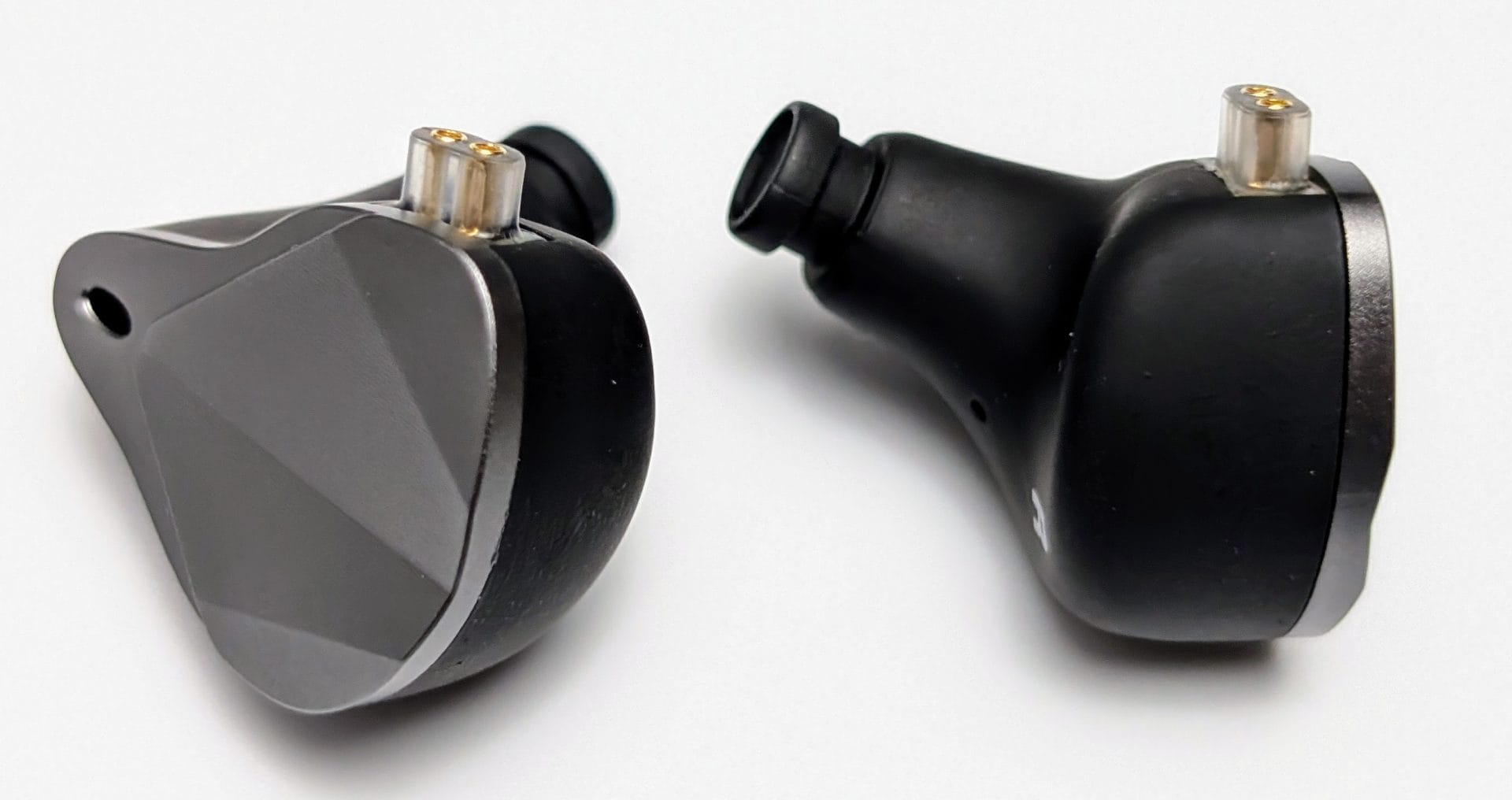 Agasound AgaOne IEMs Image showing two views of an Agasound AgaOne IEM (In-Ear Monitor). On the left, the IEM is shown with a metallic grey faceplate and gold connectors, highlighting its angular design. On the right, the IEM is shown from the back, displaying its black housing with a clear plastic connector at the top and a small hole likely for pressure relief or sound tuning.