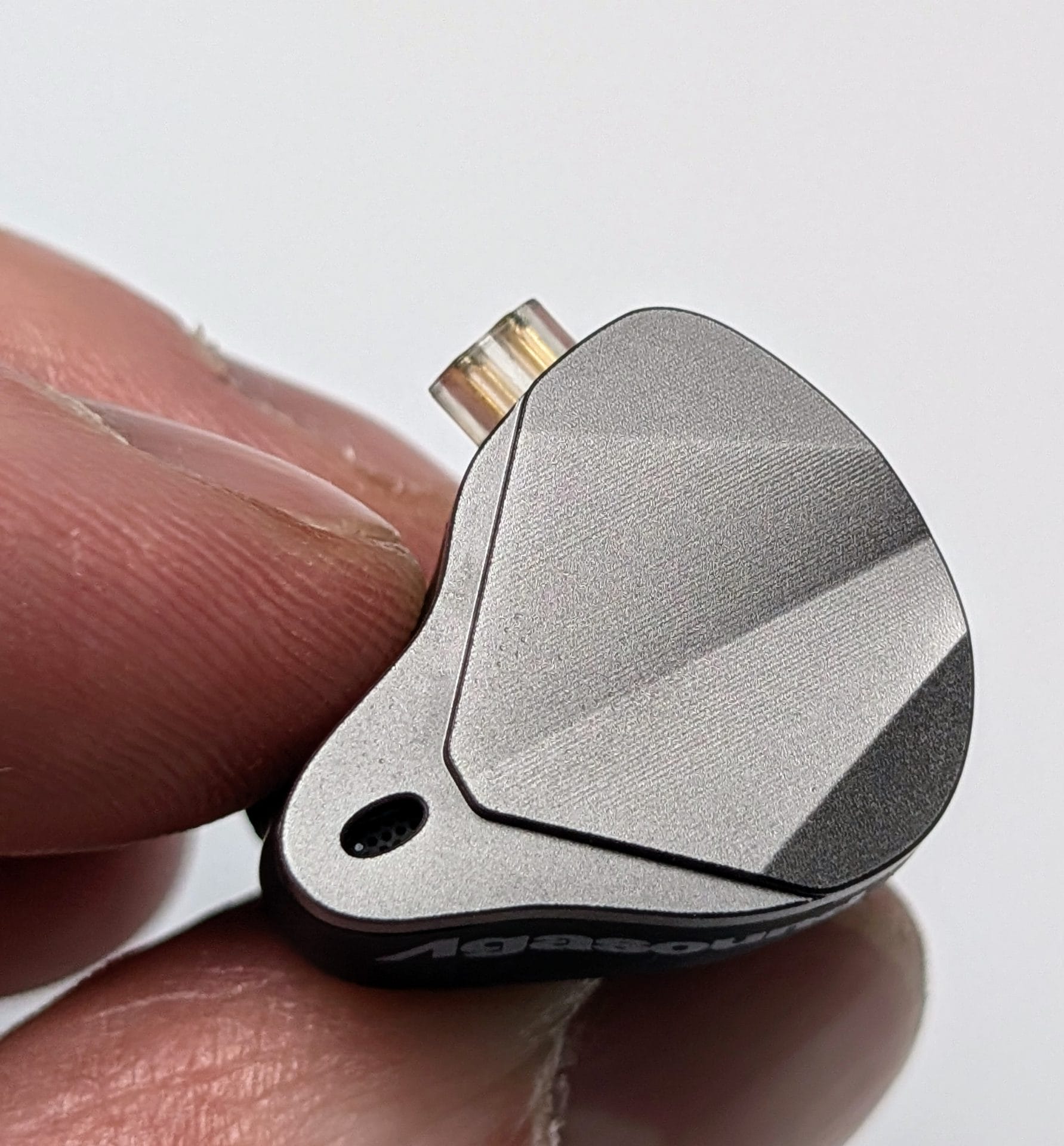 Agasound AgaOne IEMs A close-up image of an Agasound AgaOne IEM being held between two fingers. The IEM has a metallic grey faceplate with a textured finish and an angular design. There is a small hole on the faceplate, likely for sound tuning or pressure relief, and a gold connector at the top. The brand name 'Agasound' is partially visible on the black housing below the faceplate.