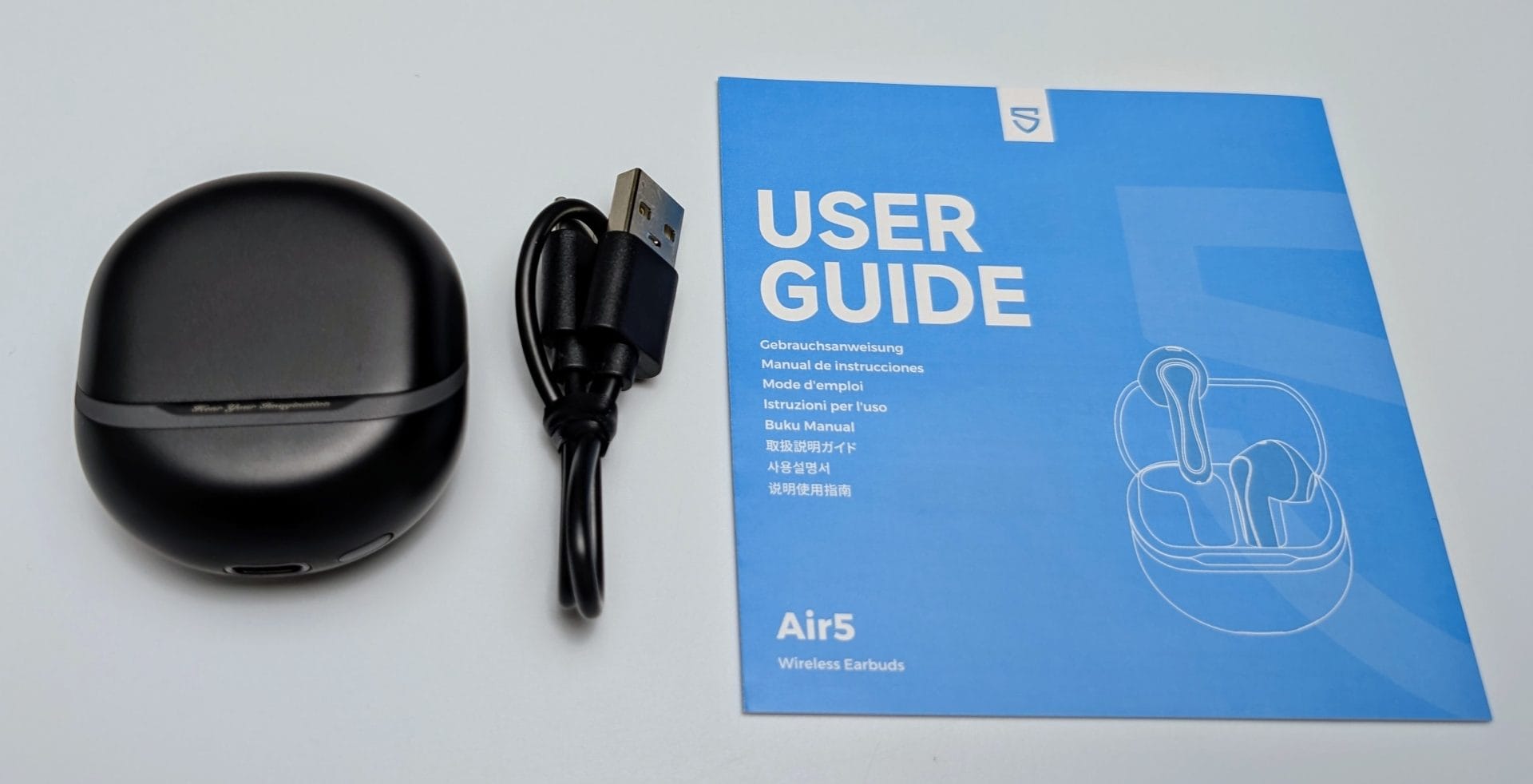 SoundPEATS Air5 Earbuds Image showing the accessories for SoundPEATS Air5 Wireless Earbuds. On the left, there is a sleek, black, oval-shaped charging case for the earbuds. Next to it, there is a black USB charging cable with a USB-A connector. On the right, there is a blue user guide with the title 'USER GUIDE' prominently displayed at the top. The guide includes translations of 'User Guide' in multiple languages such as German, Spanish, French, Italian, Japanese, Korean, and Chinese. Below the title, there's an illustration of the earbuds and their case. The model name 'Air5' and the description 'Wireless Earbuds' are printed at the bottom of the guide.