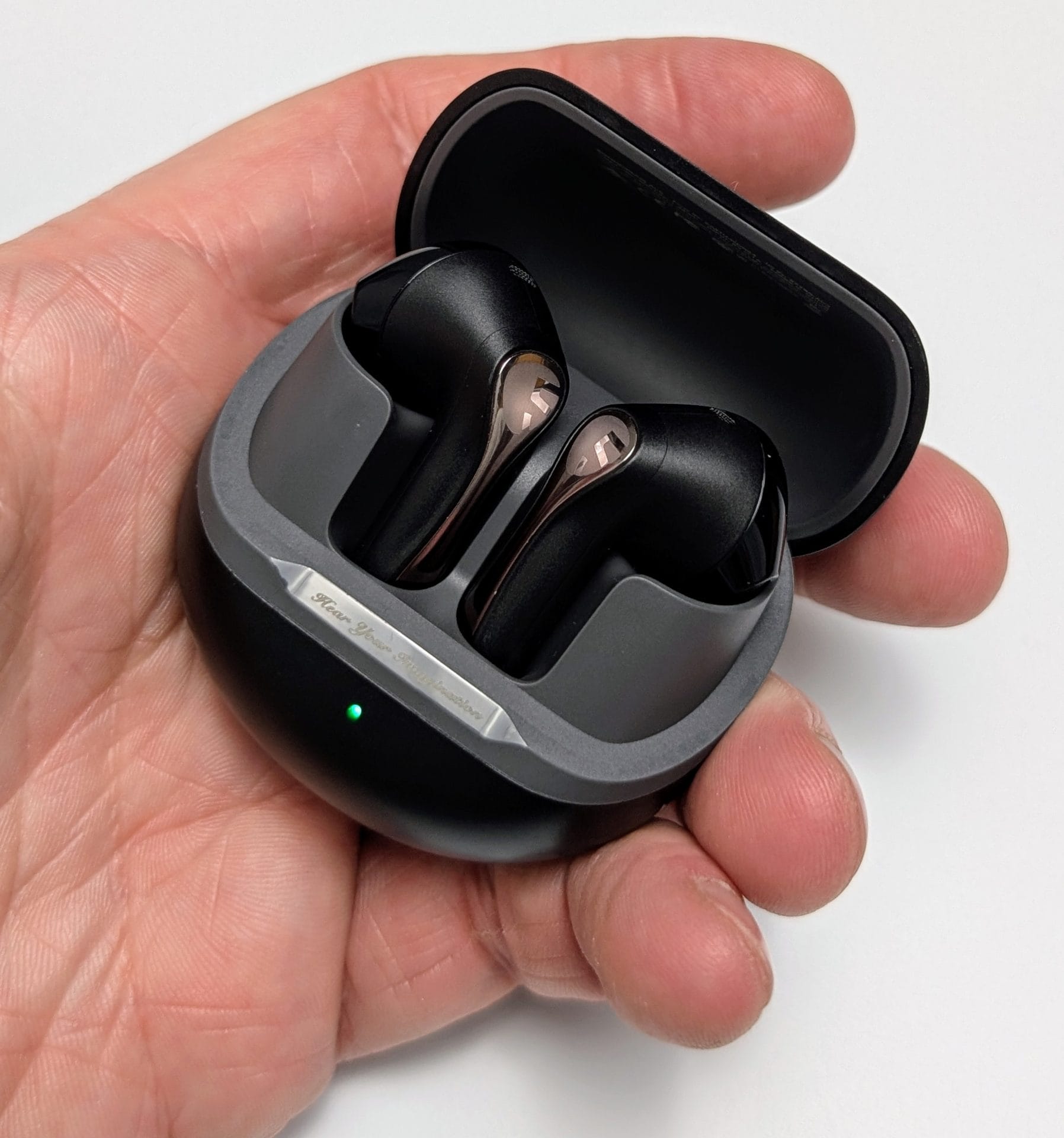 SoundPEATS Air5 Earbuds Image of a hand holding an open charging case for SoundPEATS Air5 Wireless Earbuds. The case is black with a sleek design, featuring a grey interior where the two earbuds are neatly placed. Each earbud is black with a metallic accent on the top. The case has a small green LED light indicating it is charging or fully charged. The exterior of the case has a metallic strip with the text 'For Your Imagination' engraved on it.
