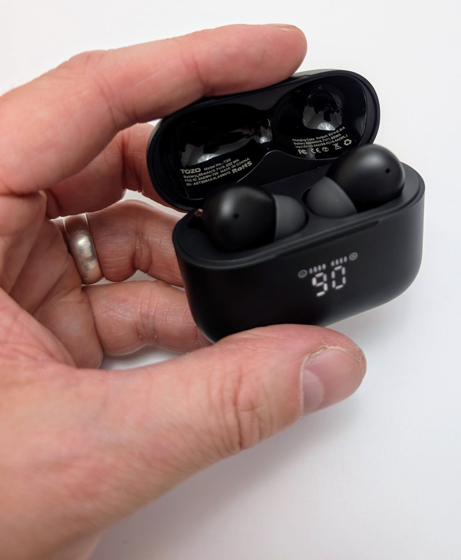 TOZO T20 Wireless Earbuds Image of a hand holding an open black TOZO T20 wireless earbuds charging case, displaying a digital battery level of 90%. Two black earbuds are inside the case, with the brand name "TOZO" visible on the interior. The hand is wearing a silver ring.