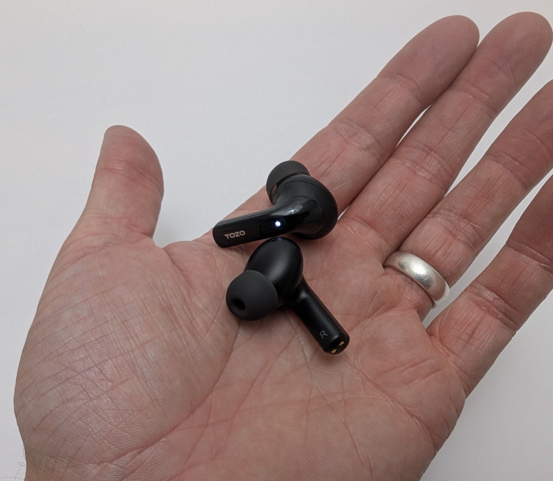 TOZO T20 Wireless Earbuds Image of a hand holding two black TOZO T20 wireless earbuds, one with a blue light indicator and labeled "L," the other labeled "R," against a plain white background. The hand is wearing a silver ring on the ring finger.