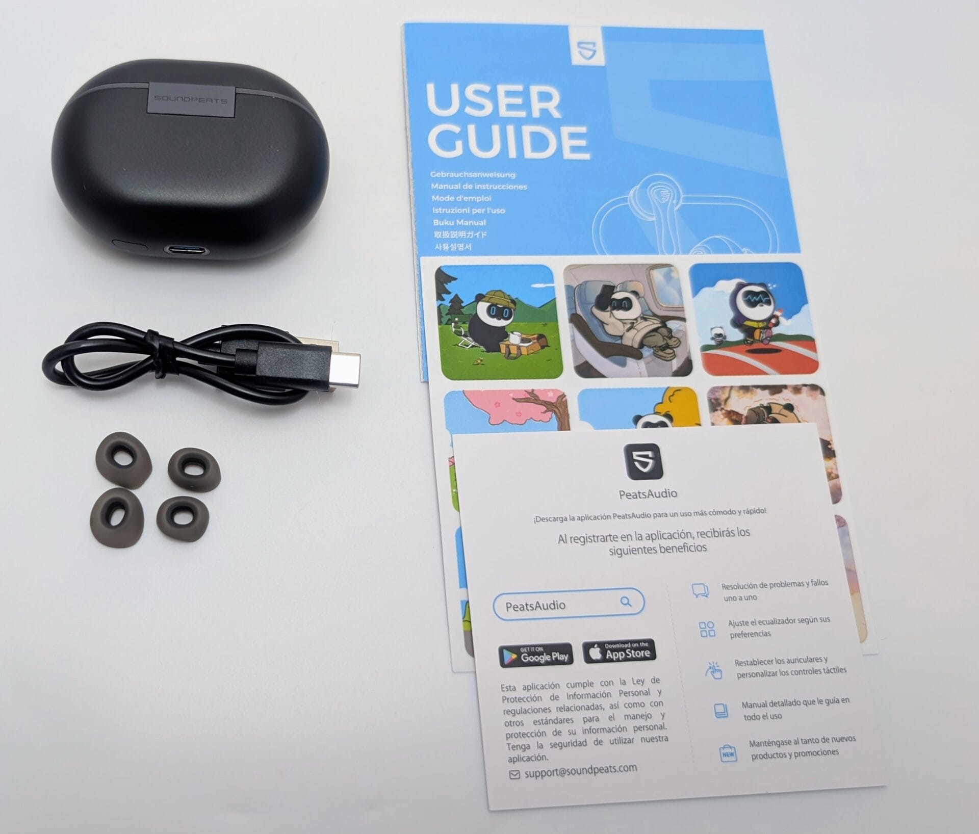 Alt text: Image of the SoundPEATS Air5 Pro earbuds unboxing, showing a black charging case with the SoundPEATS logo, a USB-C charging cable, four additional ear tips, and a user guide booklet. The user guide features a blue cover with the SoundPEATS logo, cartoon panda illustrations, and instructions in multiple languages, including details about the PeatsAudio app.