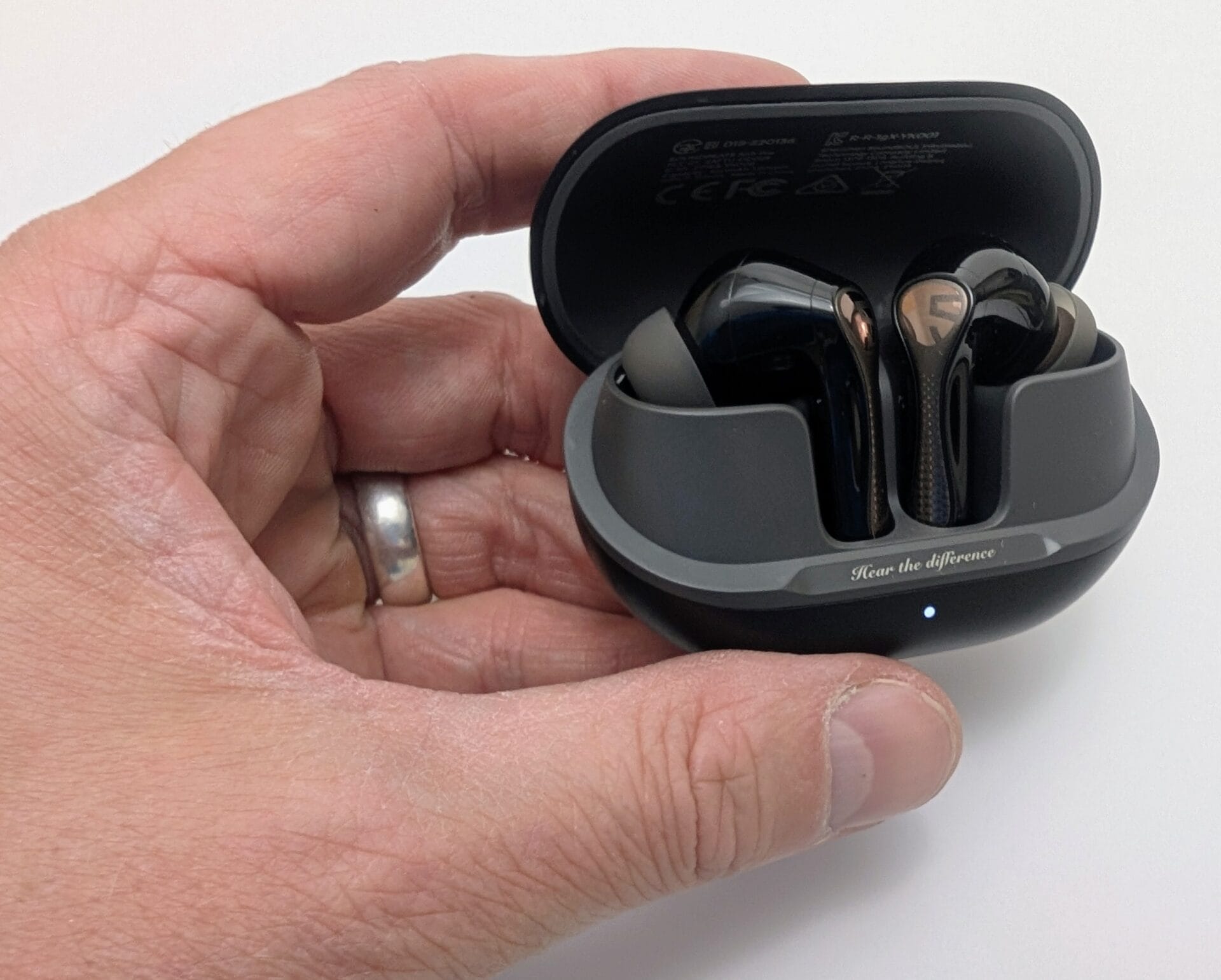 Alt text: Image of a hand holding an open SoundPEATS Air5 Pro earbuds charging case, displaying two sleek black earbuds with metallic accents inside. The case has a gray interior, the SoundPEATS logo, and the phrase "Hear the difference" embossed on it, with a small LED light visible.