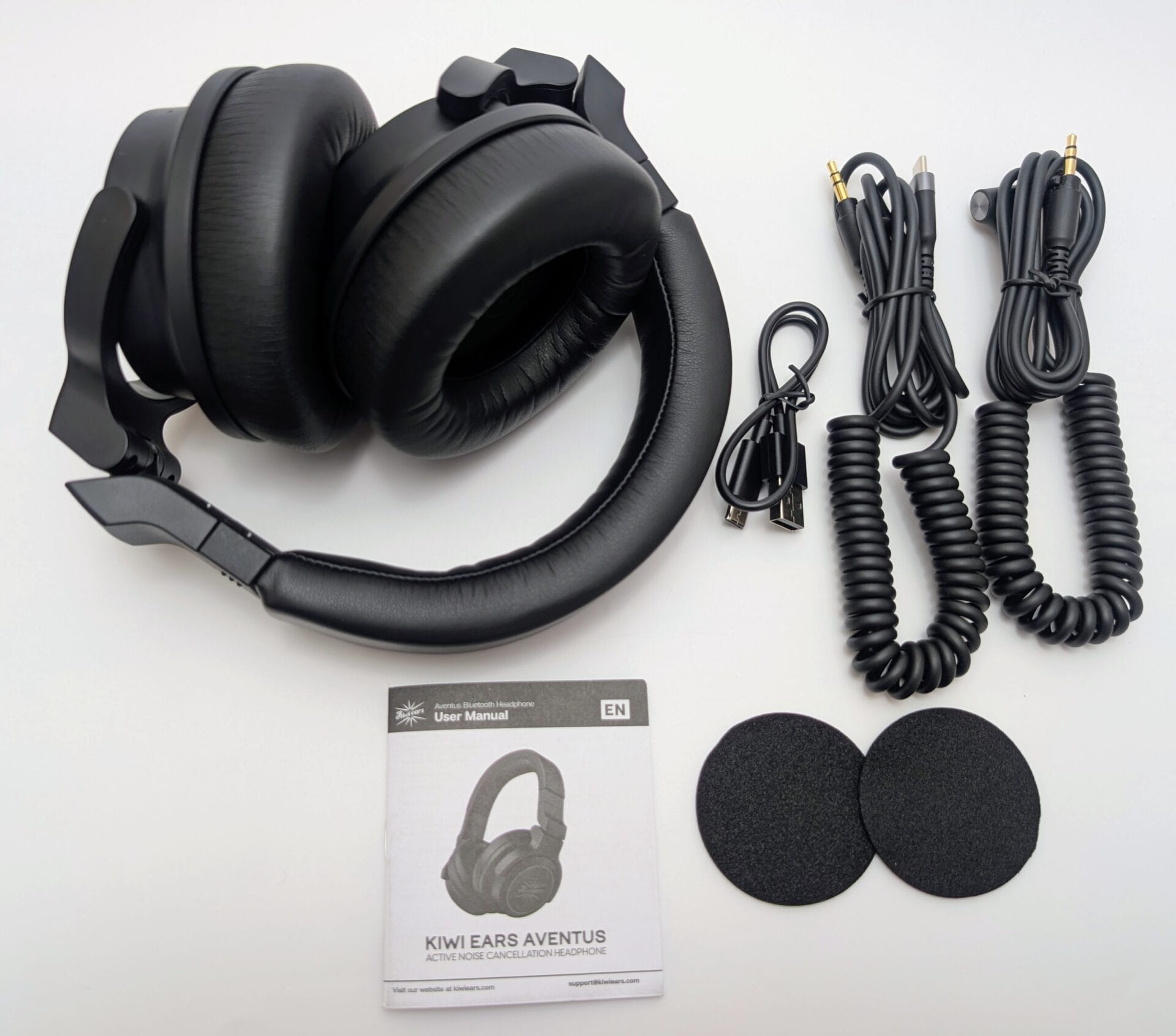 Kiwi Ears Aventus A flat lay image of the unboxed Kiwi Ears Aventus active noise-canceling headphones, featuring a black over-ear design with a cushioned headband. Included accessories are a user manual, two coiled audio cables with 3.5mm jacks, a USB cable, and two replacement ear pads, all arranged on a white background.