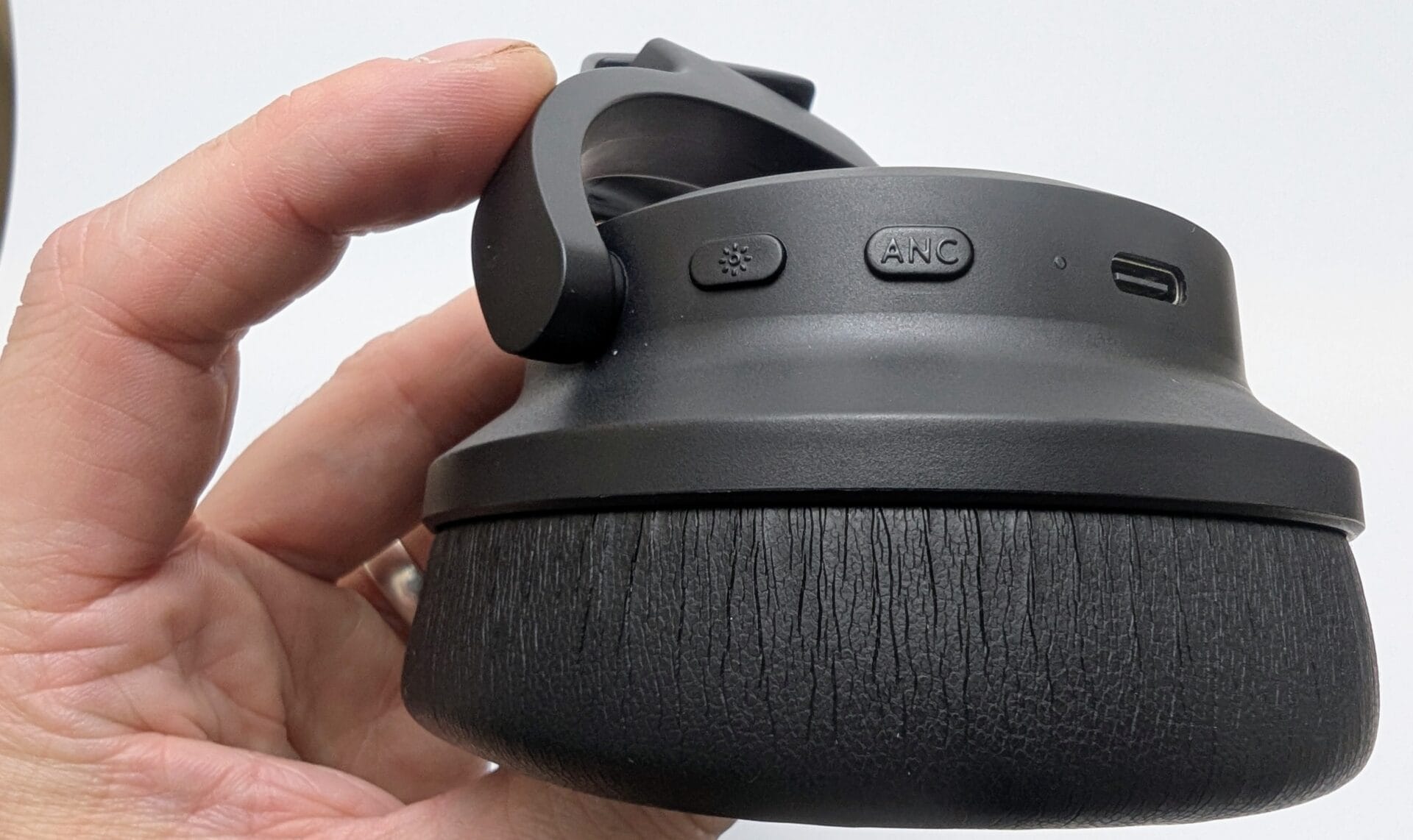 Kiwi Ears Aventus A close-up of a hand holding a black Kiwi Ears Aventus headphone ear cup, displaying the cushioned exterior, a power button with a light icon, an ANC (Active Noise Cancellation) button, and a USB-C port. The background is a plain white surface.