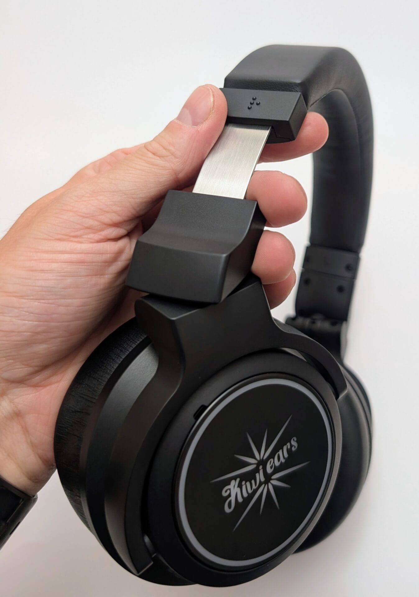 Kiwi Ears Aventus A hand holding a pair of black Kiwi Ears Aventus over-ear headphones, showcasing the cushioned headband and ear cups with the brand logo prominently displayed on the side. The background is a plain white surface.