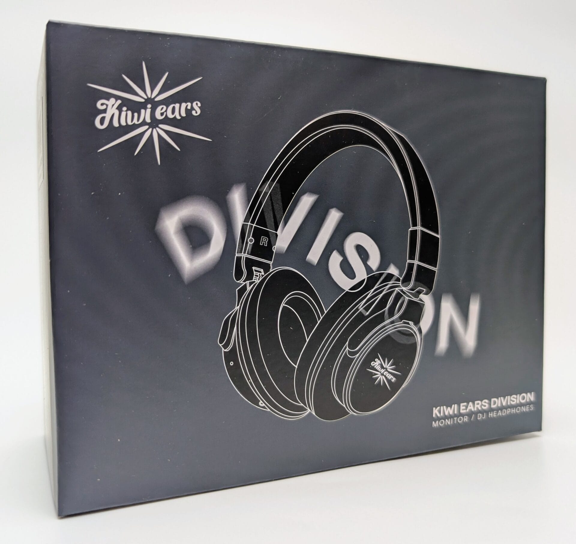 Kiwi Ears Division A close-up photograph of the Kiwi Ears Division headphone packaging box. The box has a matte black finish with a stylized white outline illustration of over-ear headphones centered on the front. The Kiwi Ears logo (a stylized starburst) appears in the top left and on the right earcup of the illustrated headphones. Large blurred text “DIVISION” is visible behind the headphones, while smaller clear text in the lower right reads “KIWI EARS DIVISION MONITOR / DJ HEADPHONES” in white. The overall design is minimalist and premium-looking.