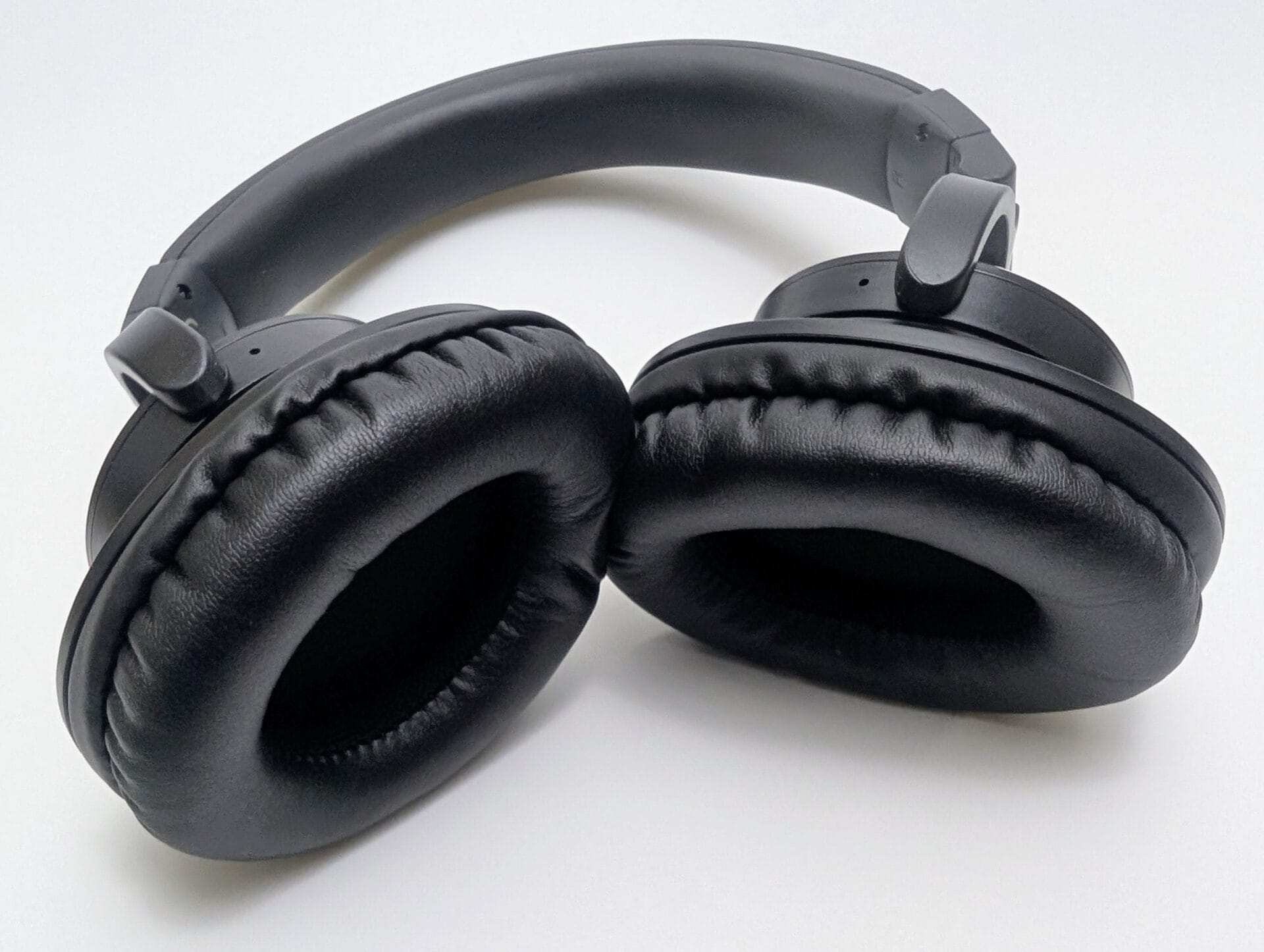 Kiwi Ears Division Close-up view of the folded Kiwi Ears Division over-ear headphones against a white background. The matte black ear cups are swiveled inward for compact storage, revealing thick, plush protein-leather earpads with visible stitching. The softly padded headband arches above, and the sturdy hinges and adjustment sliders are clearly visible, giving a premium and durable impression.