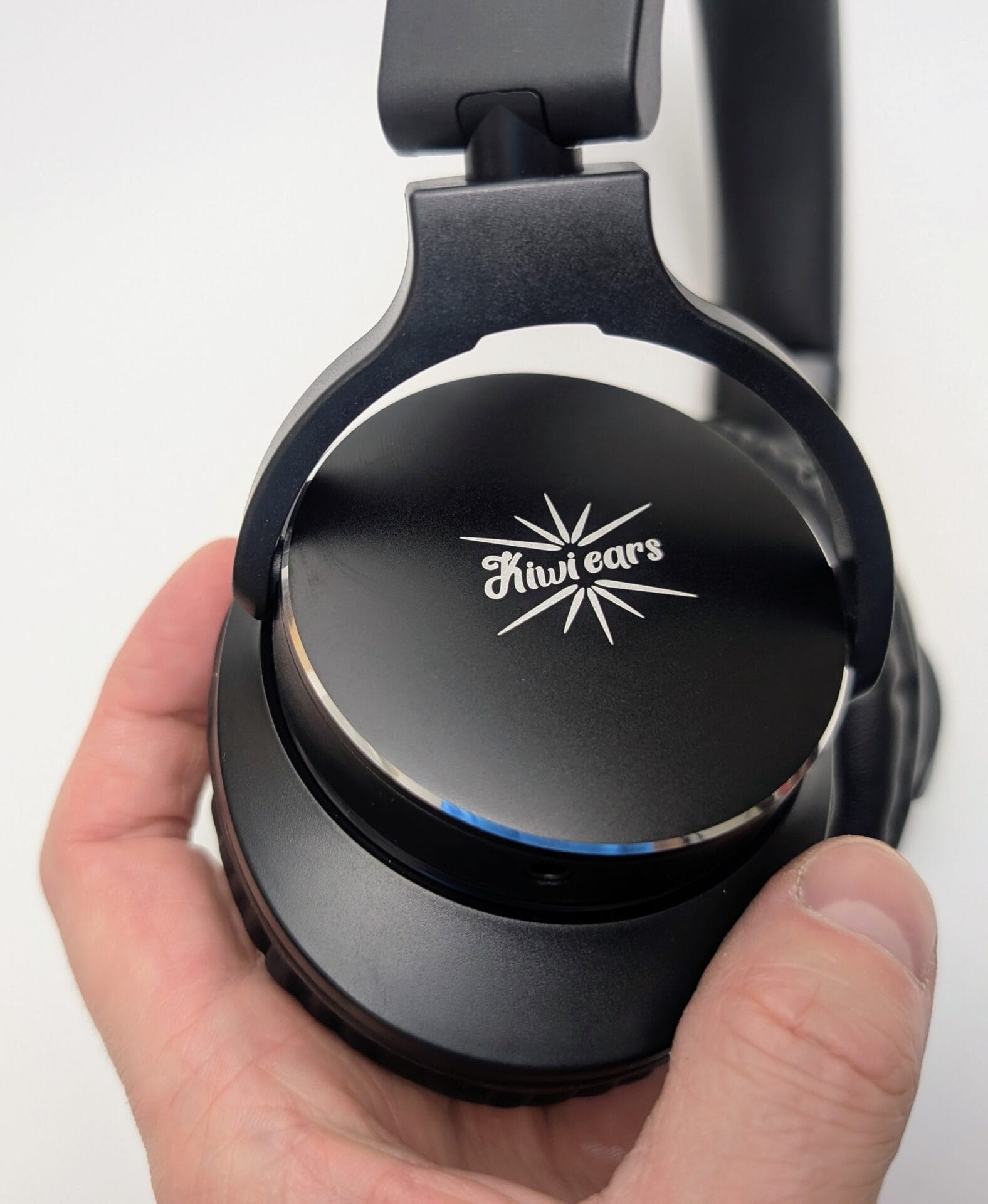 Kiwi Ears Division Close-up of the right earcup of the Kiwi Ears Division headphones held in a hand. The matte black housing features a cleanly etched silver Kiwi Ears logo (stylized starburst with the brand name in cursive script) centered on the outer shell. The foldable hinge and part of the padded headband yoke are visible, highlighting the solid metal construction and premium finish.