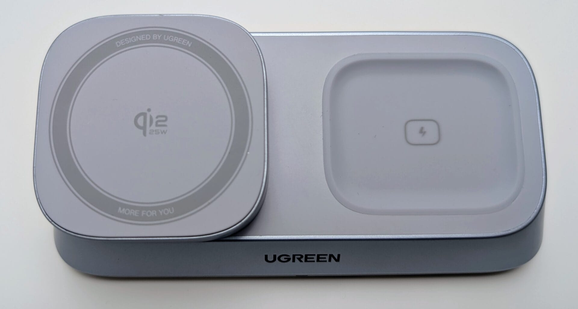 A close-up view of the UGREEN MagFlow 3-in-1 Magnetic Wireless Charger base unit. The grey/light blue charger is shown in its folded-down configuration. The left side features a round, slightly raised MagSafe-compatible charging pad labeled "Qi2 25W" with a concentric ring design. The right side is a recessed, rectangular charging area for AirPods, marked with a small lightning bolt icon. The UGREEN logo is centered on the front edge of the base.