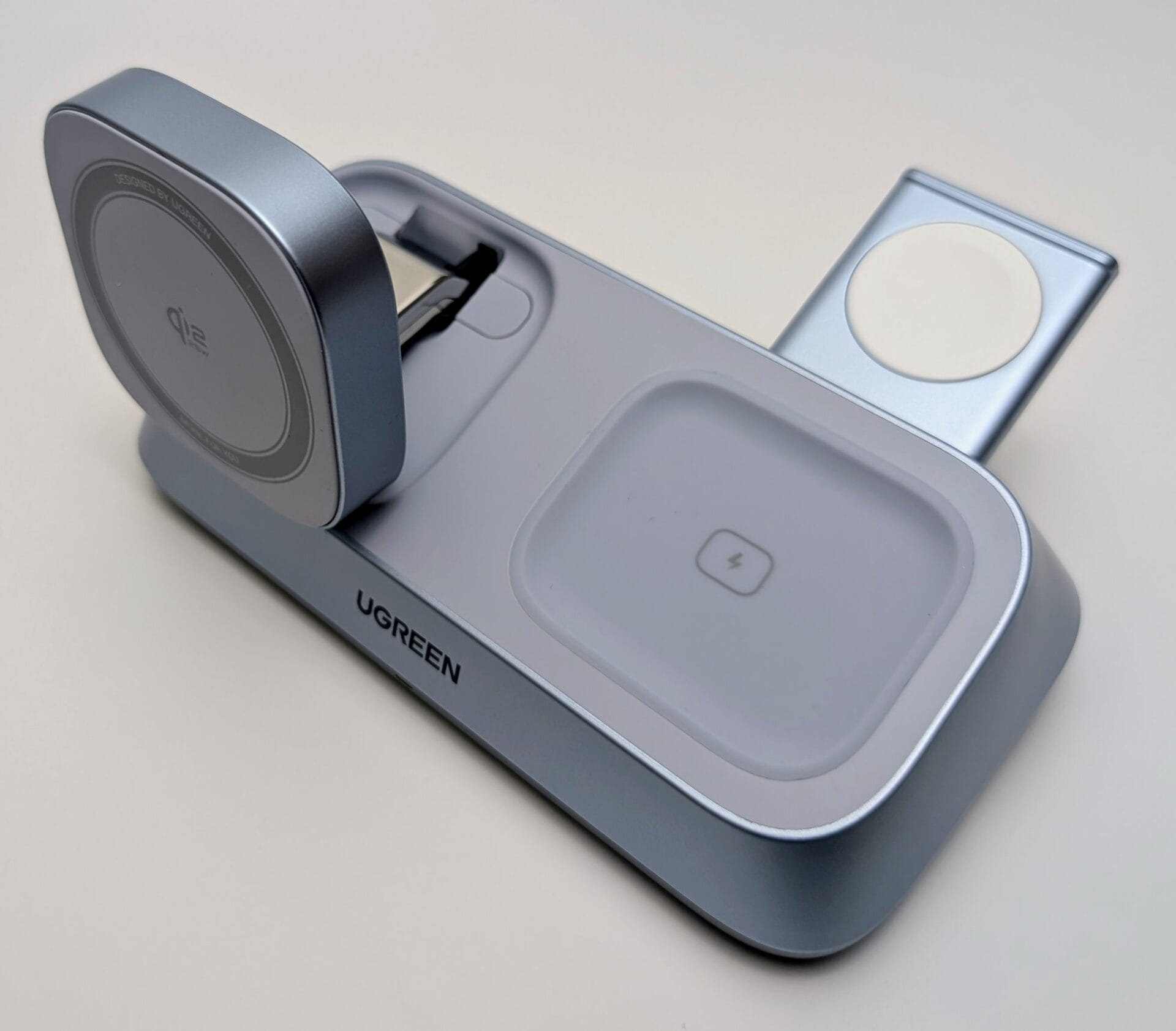 The UGREEN MagFlow 3-in-1 Magnetic Wireless Charger shown in its partially set up state. The main unit is a light blue-grey color. The round, MagSafe-compatible charging pad on the left is angled up and features the Qi2 logo. The separate Apple Watch charging arm is deployed and angled up from the right side, showing its small white magnetic charging surface. The center AirPods charging pad is flat, featuring a lightning bolt icon. The UGREEN logo is visible on the front base.