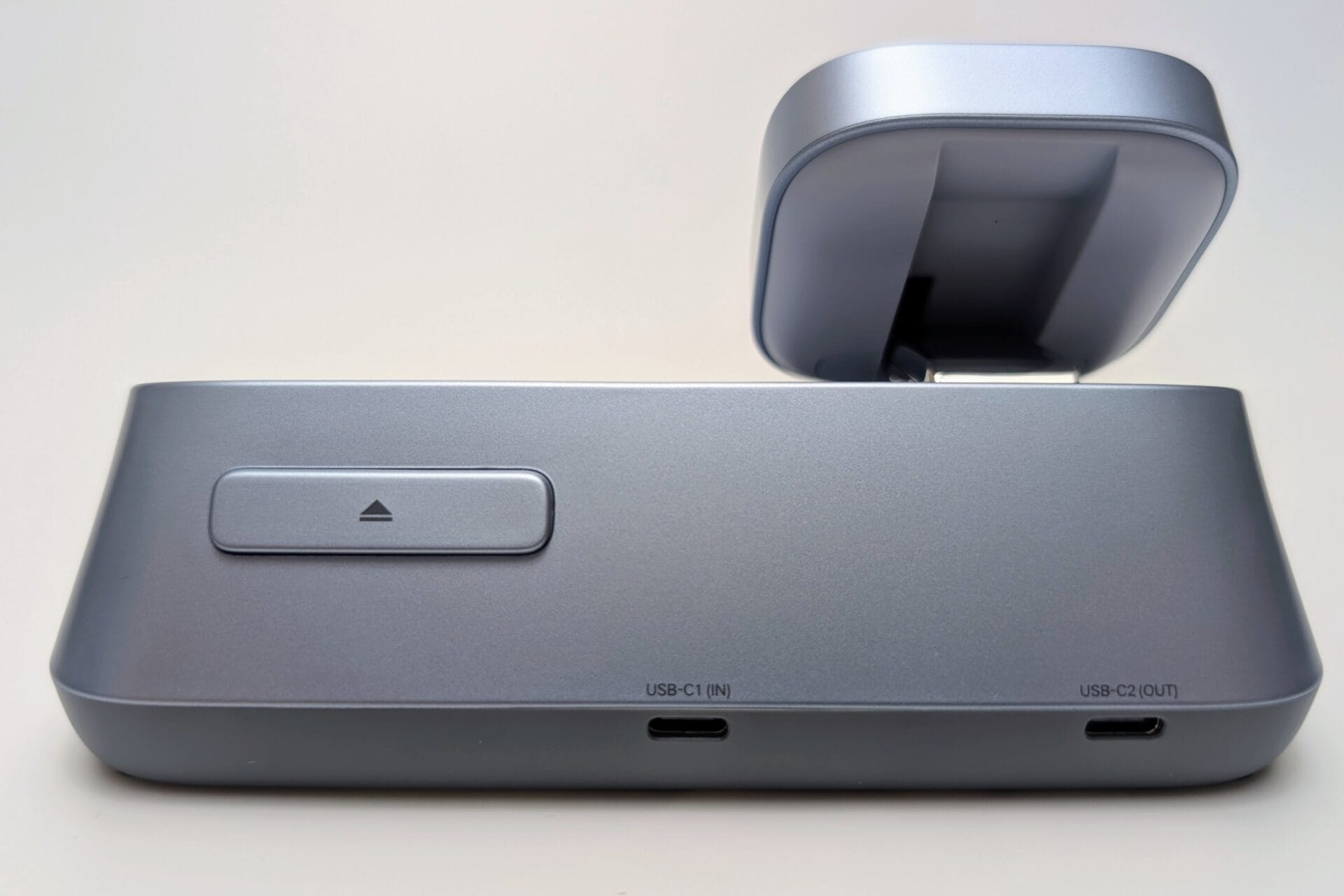 A side view of the UGREEN MagFlow 3-in-1 Magnetic Wireless Charger, focusing on the rear ports and the button for deploying the Apple Watch charger. The main unit is a smooth, light grey plastic. On the back, there is a horizontal button with a small arrow pointing up, which is used to release the Apple Watch charging arm. Below, there are two USB-C ports: one labeled "USB-C1 (IN)" for power input, and one labeled "USB-C2 (OUT)" for charging an additional device with a cable. The main MagSafe-compatible phone charger is visible in the raised position at the top.