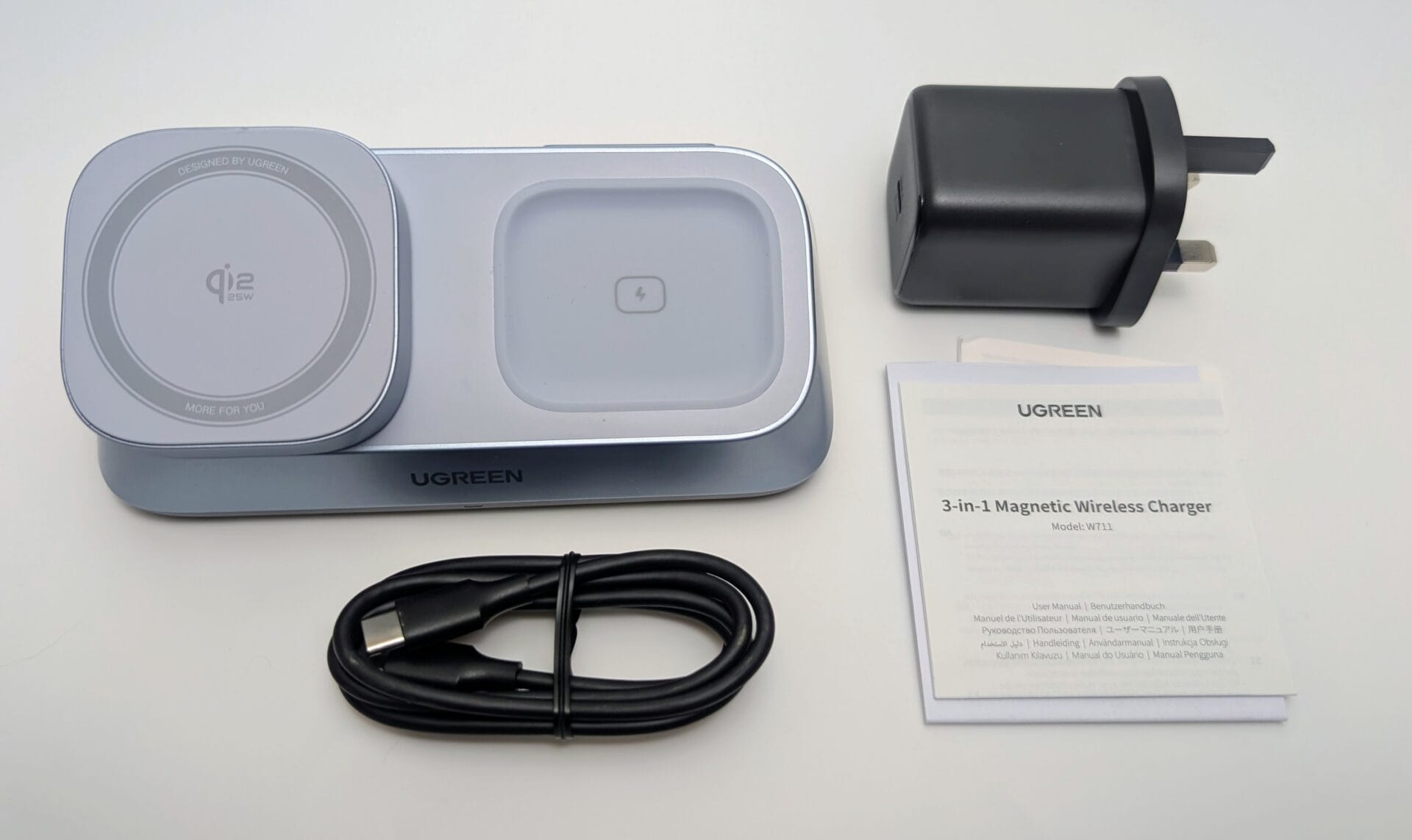 A flat lay view of the UGREEN MagFlow 3-in-1 Magnetic Wireless Charger and its included accessories, laid out on a white surface. The main charging unit is grey/light blue and features a round, foldable MagSafe-compatible charging pad on the left and a flat square charging area for AirPods on the right. Below the unit is a coiled black USB-C to USB-C cable. To the right are a black UK-style plug 45W USB-C GaN wall charger and the folded user manual titled "UGREEN 3-in-1 Magnetic Wireless Charger.