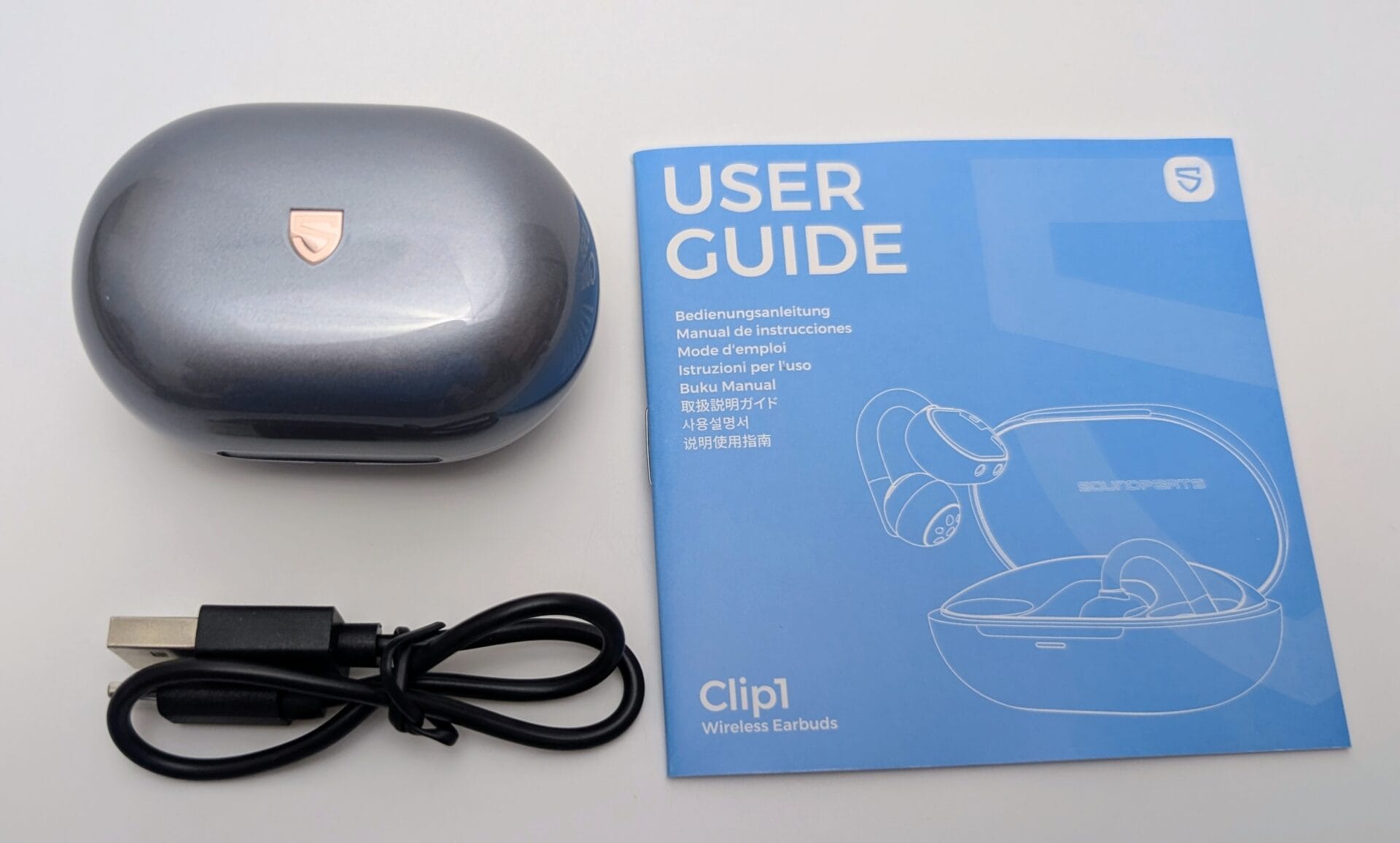 Unboxing contents of SoundPEATS Clip1 Wireless Earbuds laid out on a white background: a glossy metallic-blue charging case with rose-gold SoundPEATS logo, a black USB-A to USB-C charging cable, and a folded multilingual user guide in blue featuring illustrations of the open-ear clip design earbuds and the case. The guide prominently displays "USER GUIDE" and "Clip1 Wireless Earbuds" on the cover.