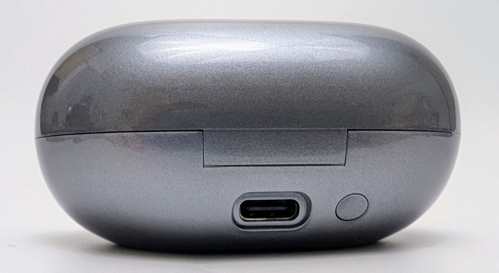 Close-up of the closed charging case for SoundPEATS Clip1 wireless earbuds in metallic gunmetal gray. The case has a glossy, rounded oval shape with a visible hinge at the front, a USB-C charging port centered on the bottom edge, and a small circular reset/pairing button to the right of the port. The finish is reflective with subtle scratches and fingerprints visible, photographed against a plain white background.