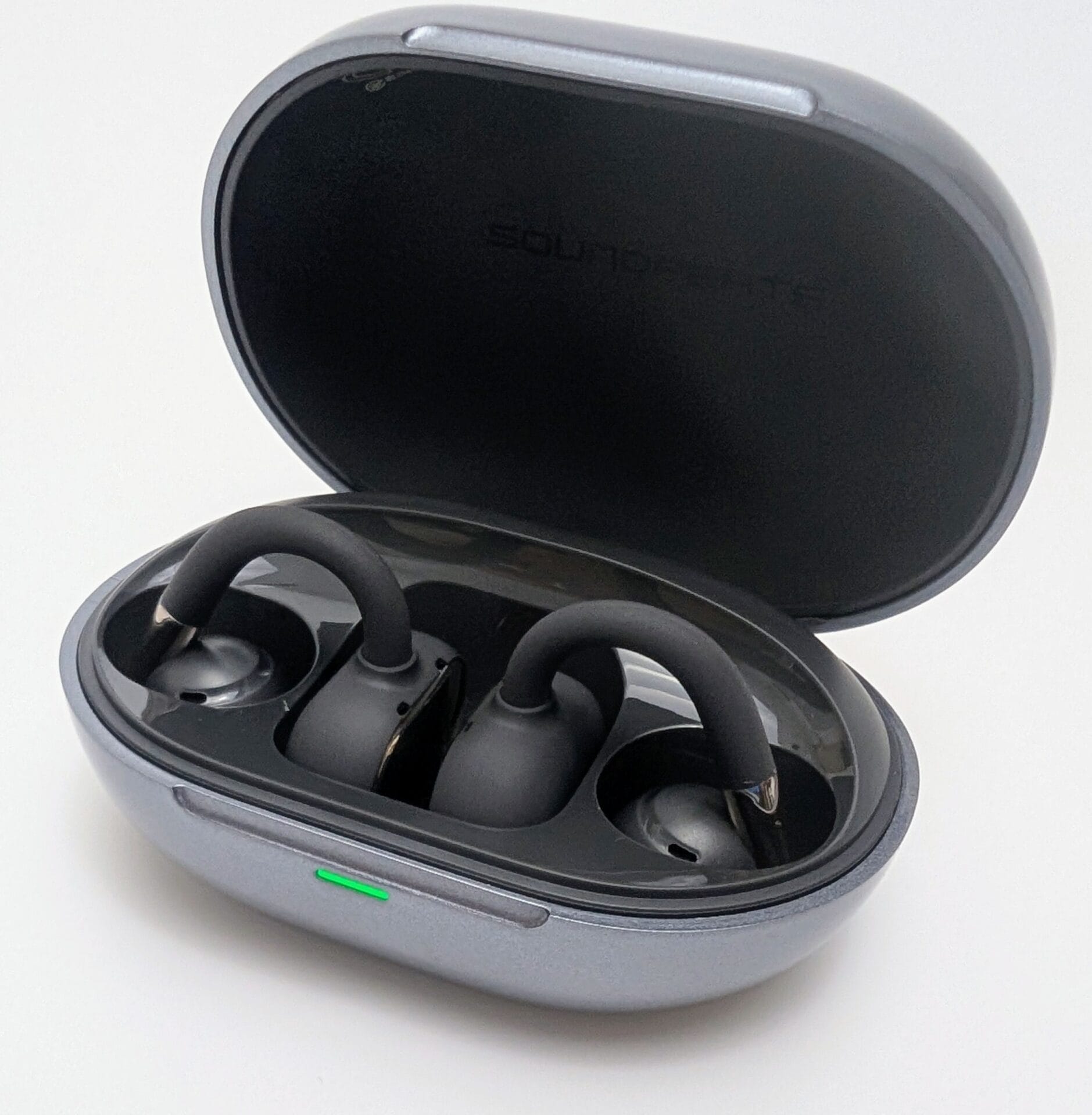 Open charging case of the SoundPEATS Clip1 wireless earbuds in metallic gunmetal gray. The lid is flipped back, revealing the two matte black open-ear clip-style earbuds securely seated in their molded slots with charging contacts aligned. A small green LED indicator glows on the front edge of the case, showing it is powered or charging. The interior lid has the faint embossed text "SOUNDPEATS" near the hinge. Photographed against a plain white background.