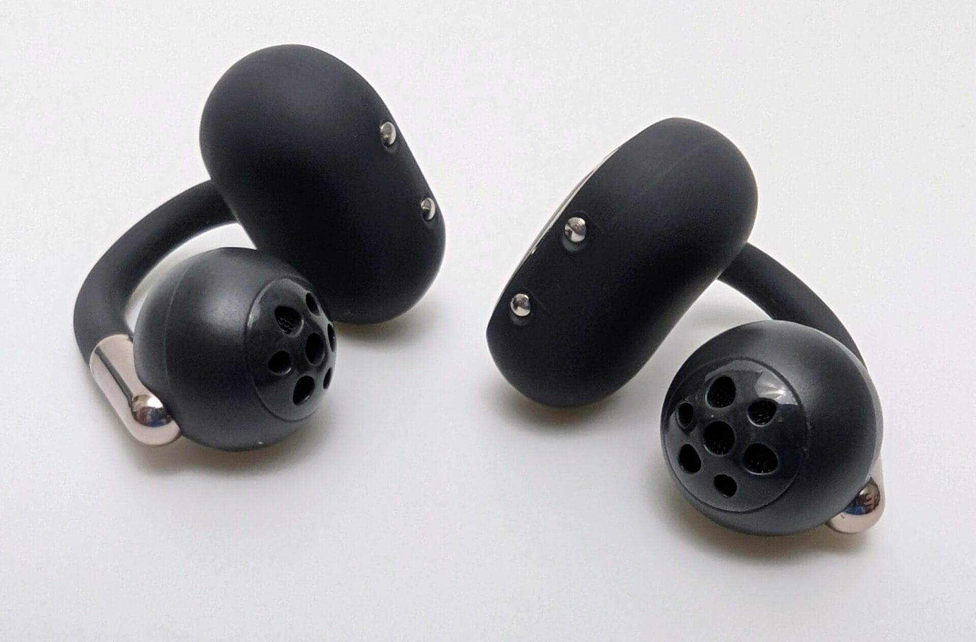 Pair of SoundPEATS Clip1 open-ear wireless earbuds in matte black, shown from multiple angles against a white background. The earbuds feature a distinctive clip-on hook design with soft rubberized ear hooks, circular speaker grilles with multiple perforations facing forward, metallic charging contacts on the inner side, and subtle silver accents near the hooks. The overall shape is ergonomic and bean-like, designed to rest outside the ear canal.