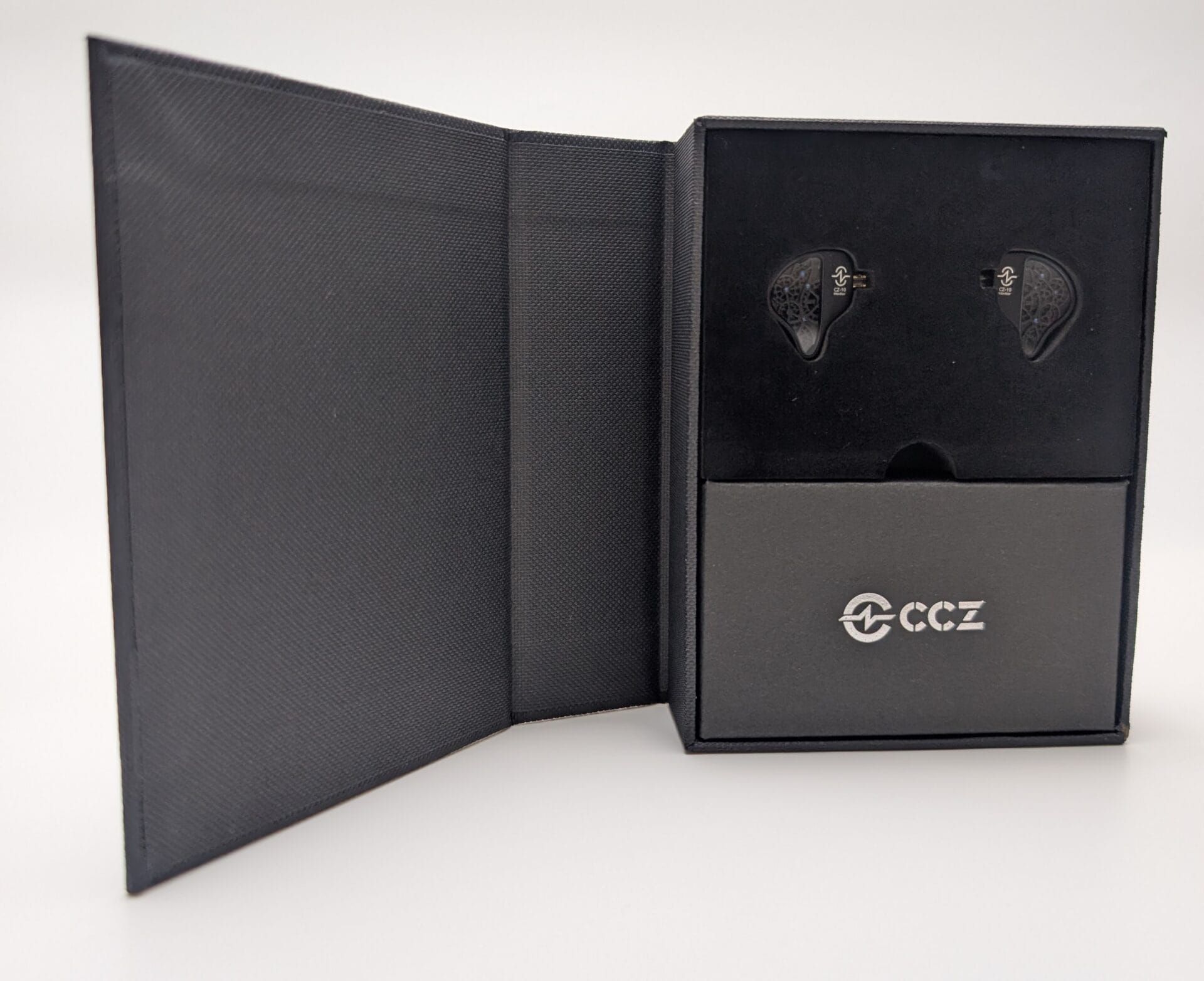 CCZ TianGong CZ10 An open, black textured presentation box for the CCZ TianGong CZ10 in-ear monitors. The two dark-colored earpieces are nestled in a black foam insert at the top, featuring an intricate, spiderweb-like pattern on their faceplates. Below them is a smaller gray accessory compartment labeled with the silver "CCZ" logo.