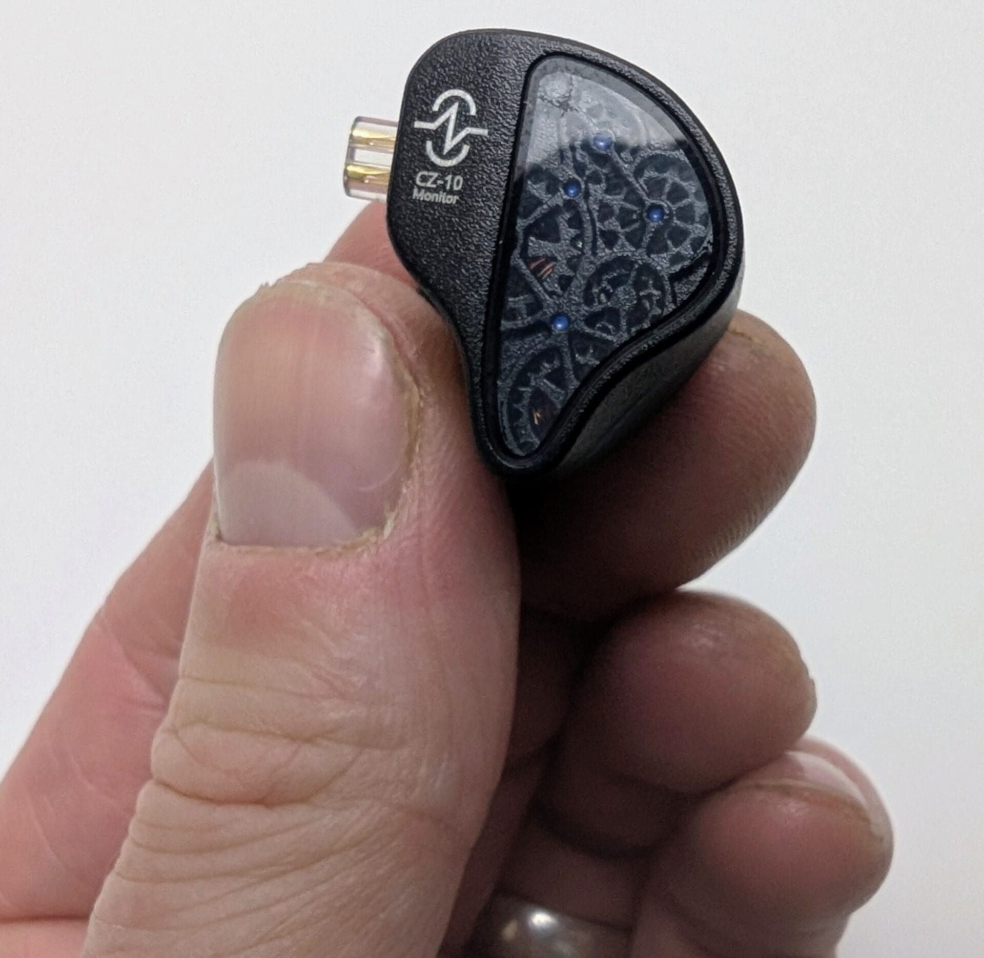 CCZ TianGong CZ10 A close-up of a person holding a CCZ TianGong CZ10 earpiece. The faceplate features a transparent window revealing a complex mechanical gear and clockwork design with small blue accents. The matte black shell includes a silver "CCZ CZ-10 Monitor" logo and a transparent 2-pin connector.