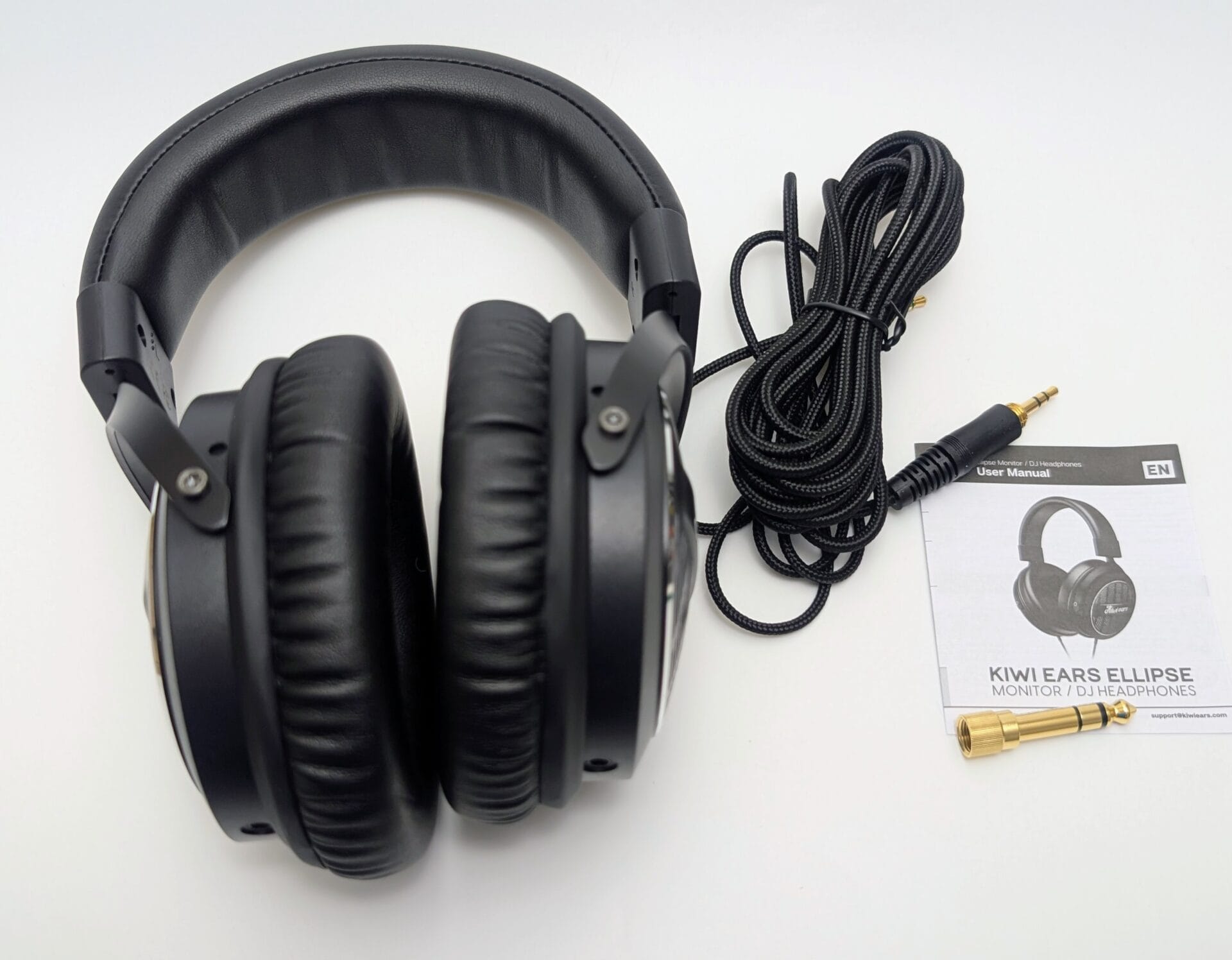 Black over-ear Kiwi Ears Ellipse DJ/monitor headphones laid flat on a white background. Features padded leatherette headband, large circular ear cups with perforated grilles and "Kiwi Ears" branding, thick black coiled cable with gold-plated 3.5mm plug, included 6.35mm adapter, and user manual booklet.