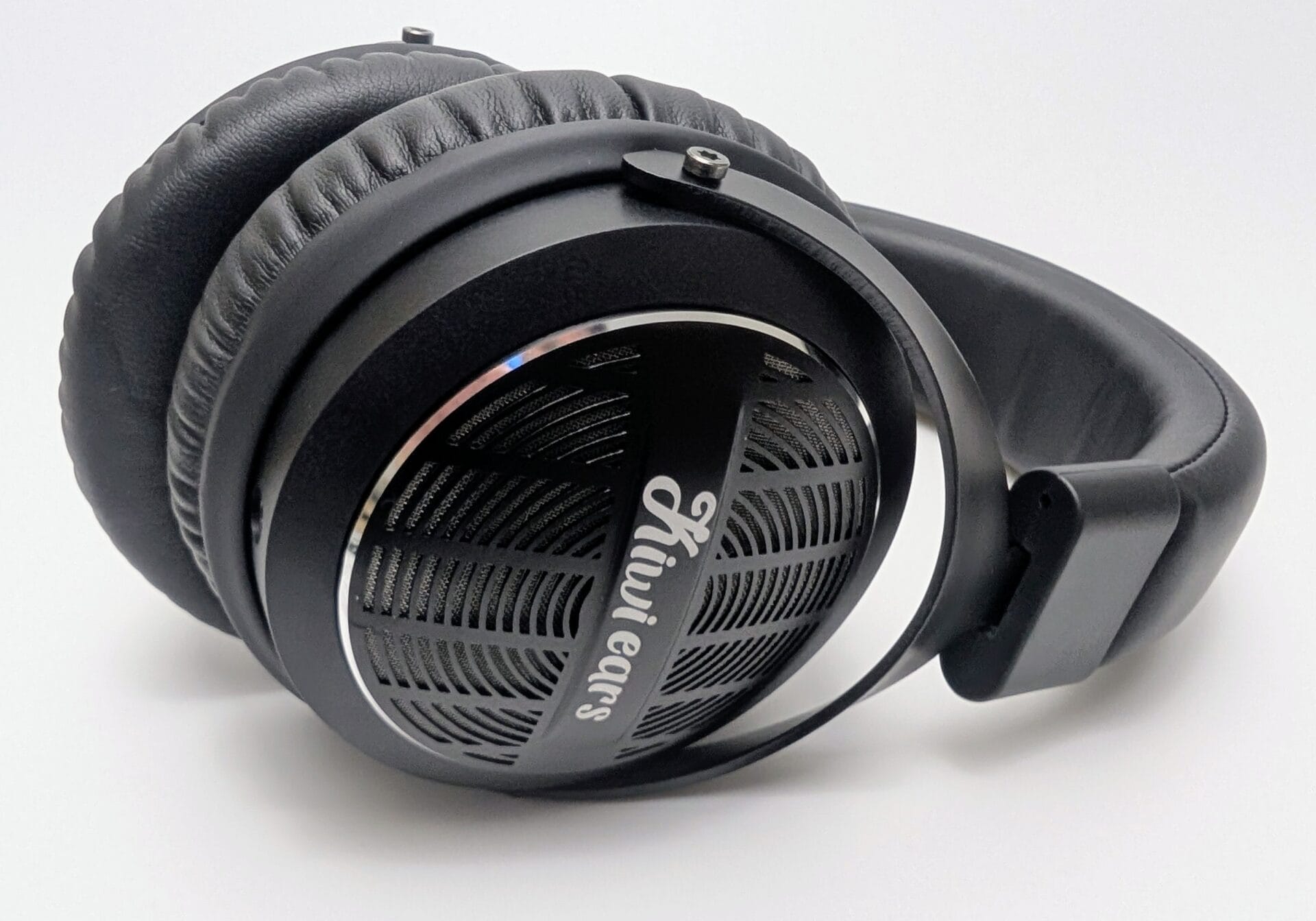 Close-up angled view of the Kiwi Ears Ellipse over-ear headphones resting on a white surface. The matte black ear cup features an open-back design with a intricate radial/spoked perforated metal grille, a shiny chrome accent ring, and the brand name “Kiwi Ears” in elegant white script across the center. Thick, padded black leatherette ear pads are visible, along with part of the matching black headband and adjustable yoke with a small exposed screw. Sleek, premium DJ/monitor style with subtle iridescent sheen on the chrome ring.