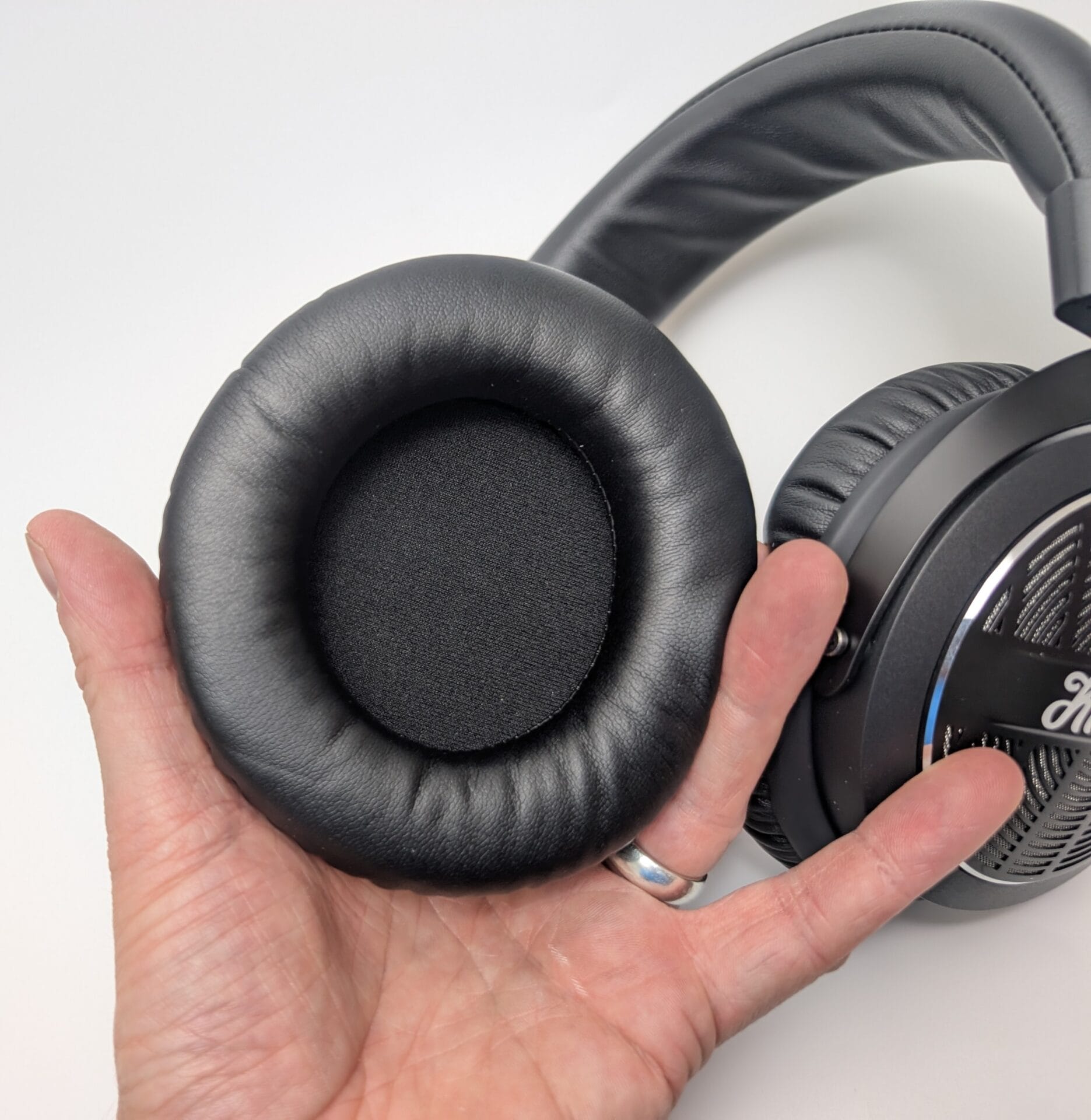 Close-up of the ear pad on the Kiwi Ears Ellipse over-ear headphones. The large, round ear cushion is covered in soft black leatherette with a slightly textured, pebbled finish. The inner opening is circular and deep, showing a dark fabric driver cover inside. The headphone's matte black outer ear cup is visible in the background, featuring a perforated metallic grille with radial pattern, chrome accent ring, and subtle "Kiwi Ears" branding. A hand is holding the headphone, with a silver ring visible on one finger.