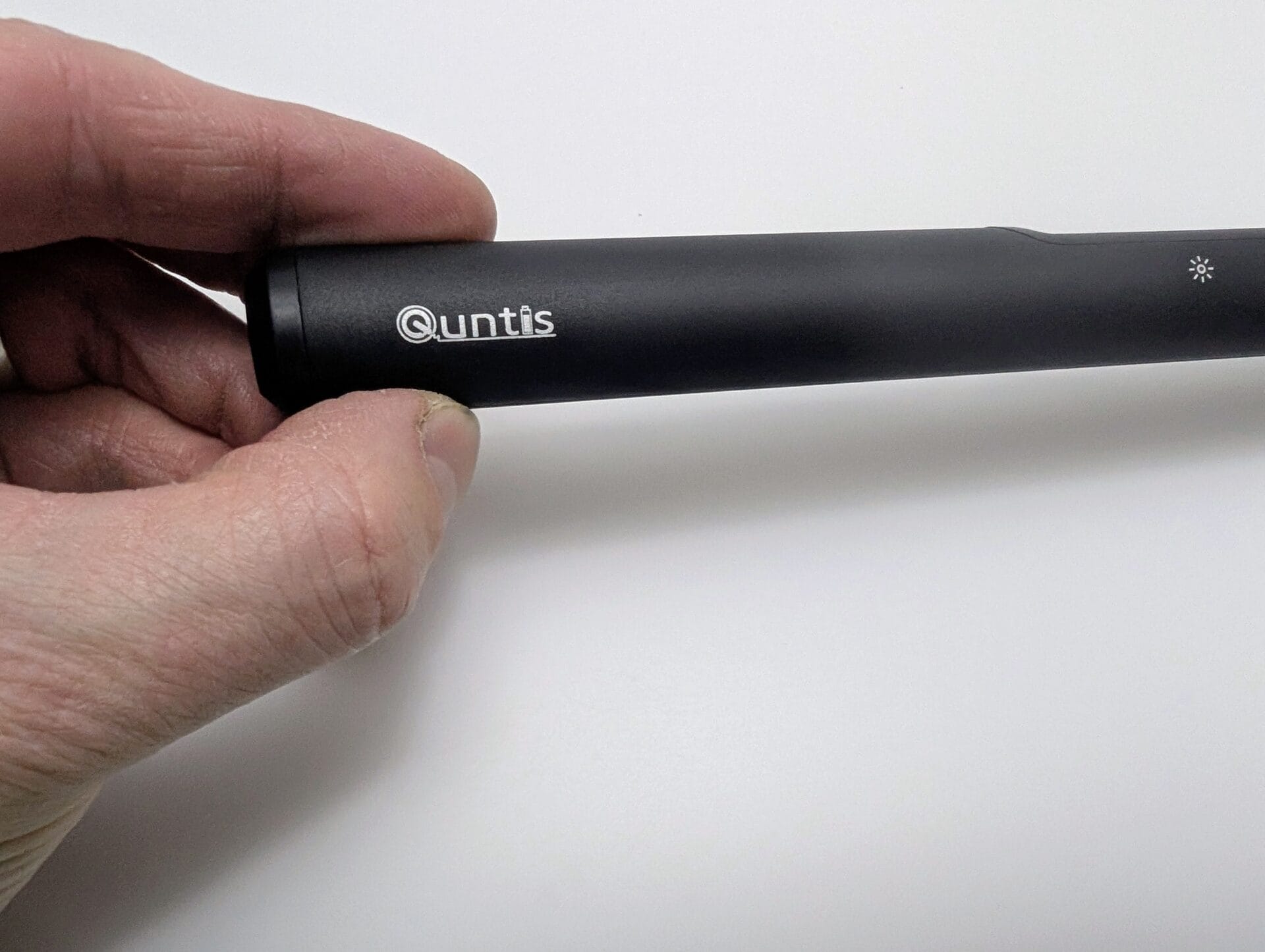 Hand holding the end of the black Quntis LED monitor light bar, displaying the 'Quntis' logo and brightness/sun icon on the matte black cylindrical body.