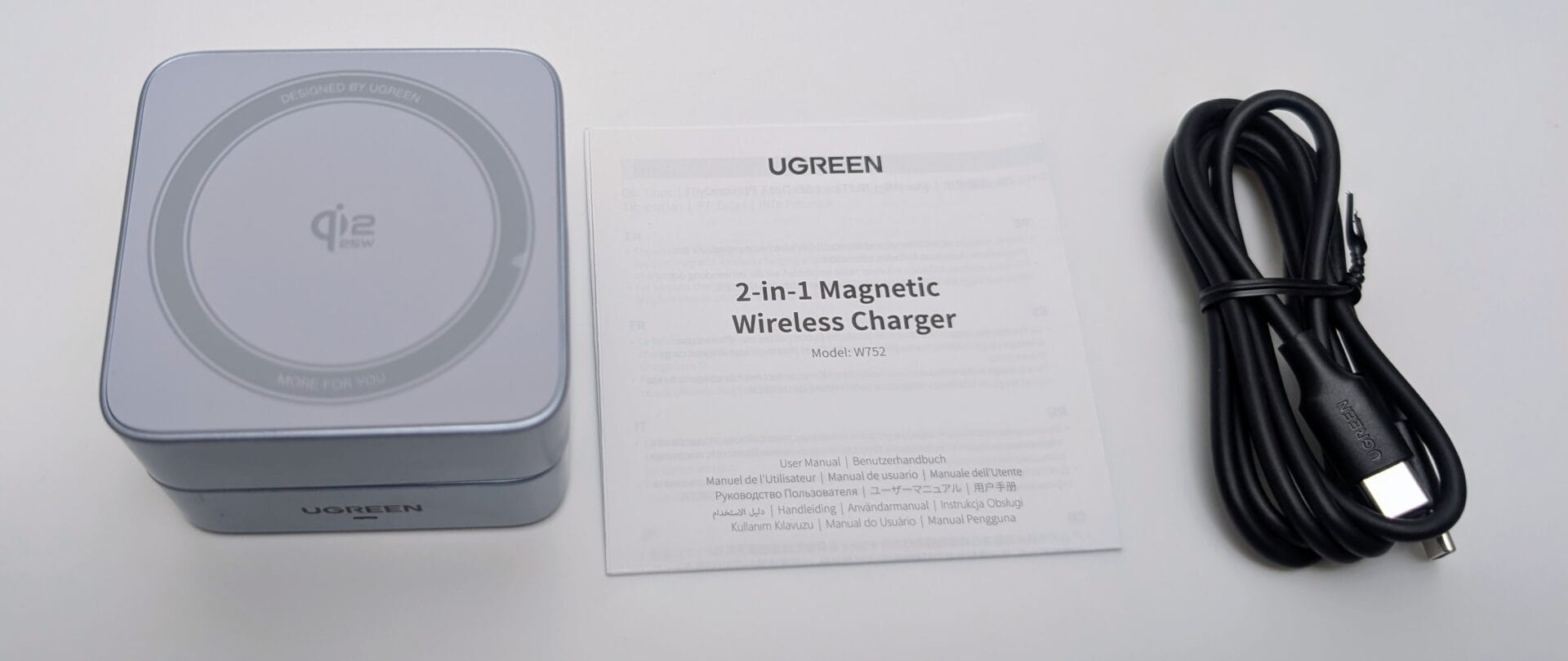 UGREEN MagFlow 2-in-1 Magnetic Wireless Charger 25W The unboxed UGREEN MagFlow 2-in-1 Magnetic Wireless Charger (25W, Qi2-certified, model W752) on a white background, showing the compact square silver-gray base with a flat-folded metallic arm and circular Qi2 charging pad labeled "Qi2 25W", "DESIGNED BY UGREEN", and "MORE FOR YOU". Next to it is the open multi-language user manual displaying the title "2-in-1 Magnetic Wireless Charger" and model details, plus a coiled black USB-C to USB-C cable with UGREEN branding.