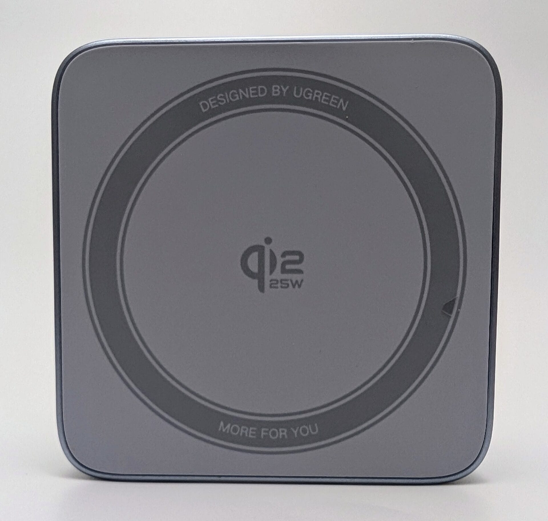 Charging Area – Image Source – My Helpful Hints (Subject to Copyright) The top view of the UGREEN MagFlow 2-in-1 Magnetic Wireless Charger (25W, Qi2-certified, model W752) in its folded, portable form. It is a compact square device with a matte silver-gray aluminum-like finish and rounded edges. The center features a large circular Qi2 charging pad with concentric gray rings, prominently displaying the "Qi2" logo in the middle surrounded by "25W". Around the outer edge of the pad are embossed texts: "DESIGNED BY UGREEN" at the top arc and "MORE FOR YOU" at the bottom arc. The overall design is minimalist and premium, shown against a plain white background.