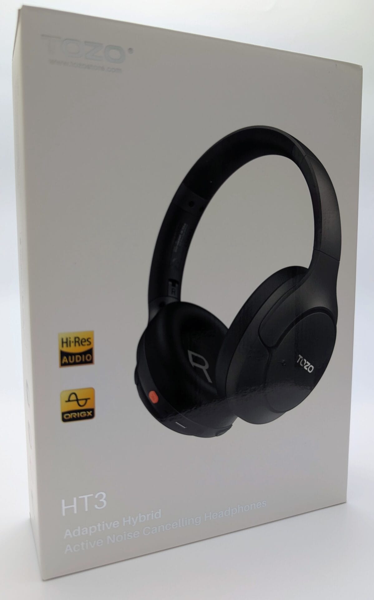 The image shows an unboxed TOZO HT3 Adaptive Hybrid Active Noise Cancelling Headphones set laid out on a light surface. The matte black over-ear headphones are prominently displayed in the center, folded slightly with the padded headband curved upward. The large, circular ear cups feature the white "TOZO" logo embossed on both sides, a small orange accent button and microphone grille on the right ear cup, and subtle perforations on the left. A matching black USB-C charging cable (with USB-A to USB-C connectors) lies coiled nearby, secured with a black tie. Accompanying the headphones are two folded paper documents: a "Quick Guide" card with a simple line drawing of the headphones, and a thicker "User Manual" booklet labeled "HT3 Adaptive Hybrid Active Noise Cancelling Headphones" with the TOZO logo and website (www.tozostore.com) (www.tozostore.com). The overall layout is clean and organized, highlighting the product's minimalist design, included accessories, and documentation.