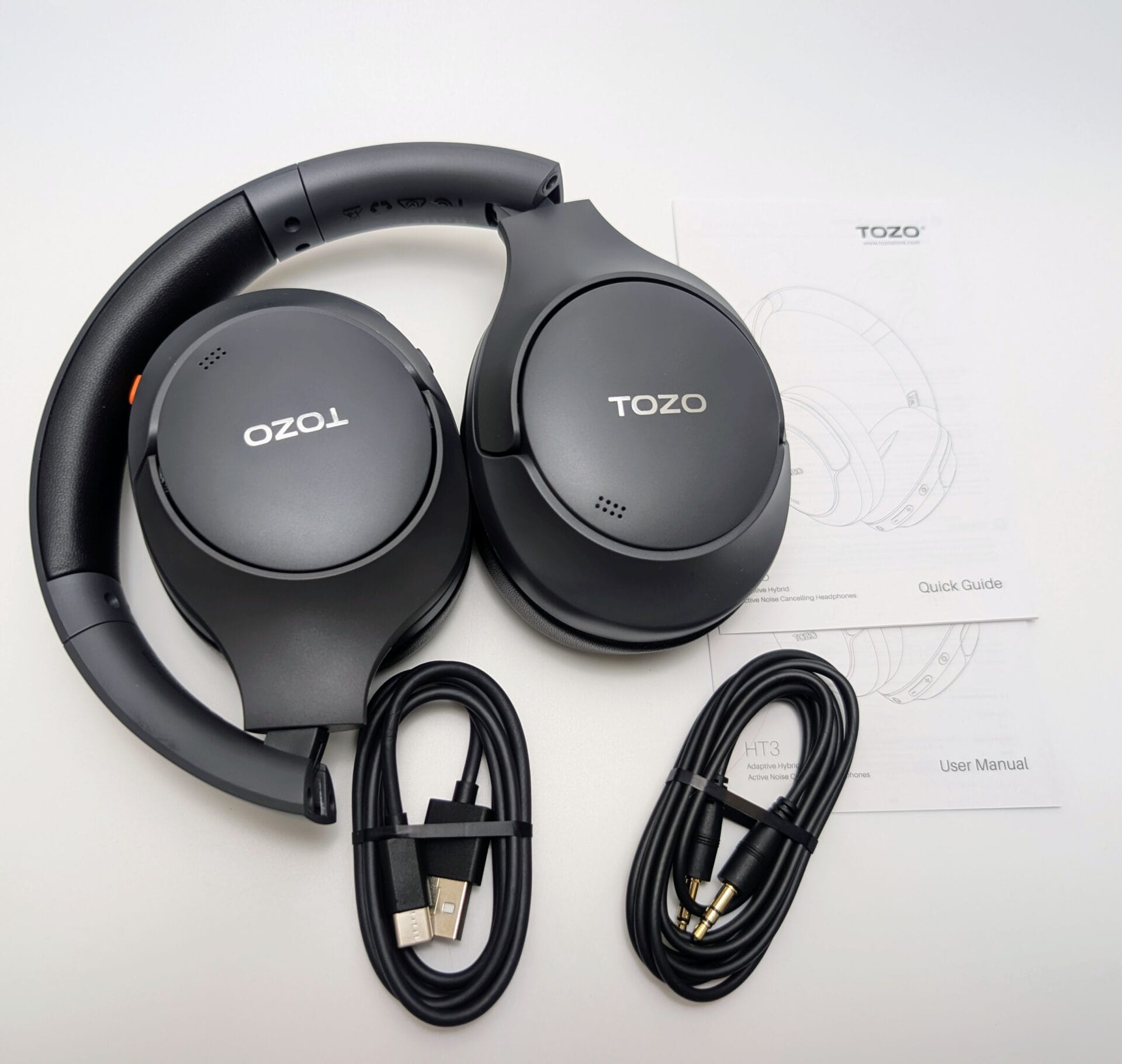 The image shows an unboxed TOZO HT3 Adaptive Hybrid Active Noise Cancelling Headphones set laid out on a light surface. The matte black over-ear headphones are prominently displayed in the center, folded slightly with the padded headband curved upward. The large, circular ear cups feature the white "TOZO" logo embossed on both sides, a small orange accent button and microphone grille on the right ear cup, and subtle perforations on the left. A matching black USB-C charging cable (with USB-A to USB-C connectors) lies coiled nearby, secured with a black tie. Accompanying the headphones are two folded paper documents: a "Quick Guide" card with a simple line drawing of the headphones, and a thicker "User Manual" booklet labeled "HT3 Adaptive Hybrid Active Noise Cancelling Headphones" with the TOZO logo and website (www.tozostore.com) (www.tozostore.com). The overall layout is clean and organized, highlighting the product's minimalist design, included accessories, and documentation.