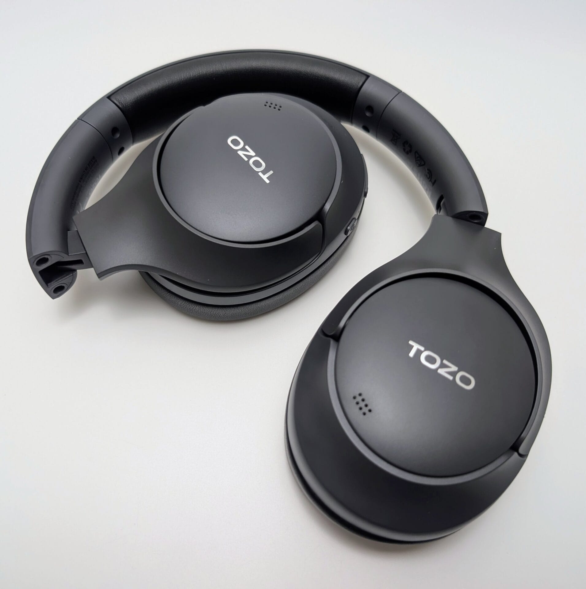 The image shows a top-down angled view of the TOZO HT3 Adaptive Hybrid Active Noise Cancelling Headphones placed on a light surface in matte black. The over-ear headphones are positioned with one side slightly elevated, revealing the padded, adjustable headband with a textured leather-like finish and small ventilation holes near the hinges. The large, circular ear cups feature flat, smooth matte outer panels prominently displaying the white "TOZO" logo in uppercase letters centered on each. Small clusters of perforations (likely microphone arrays or vents) are visible near the top of both ear cups. The ear pads are thick, cushioned, and covered in matching black textured protein leather, contributing to a comfortable, premium-looking over-ear design. The overall aesthetic is sleek and minimalist, with subtle hinges allowing the ear cups to fold inward for portability, emphasizing the headphones' ergonomic build, long battery life potential, and modern wireless ANC features.