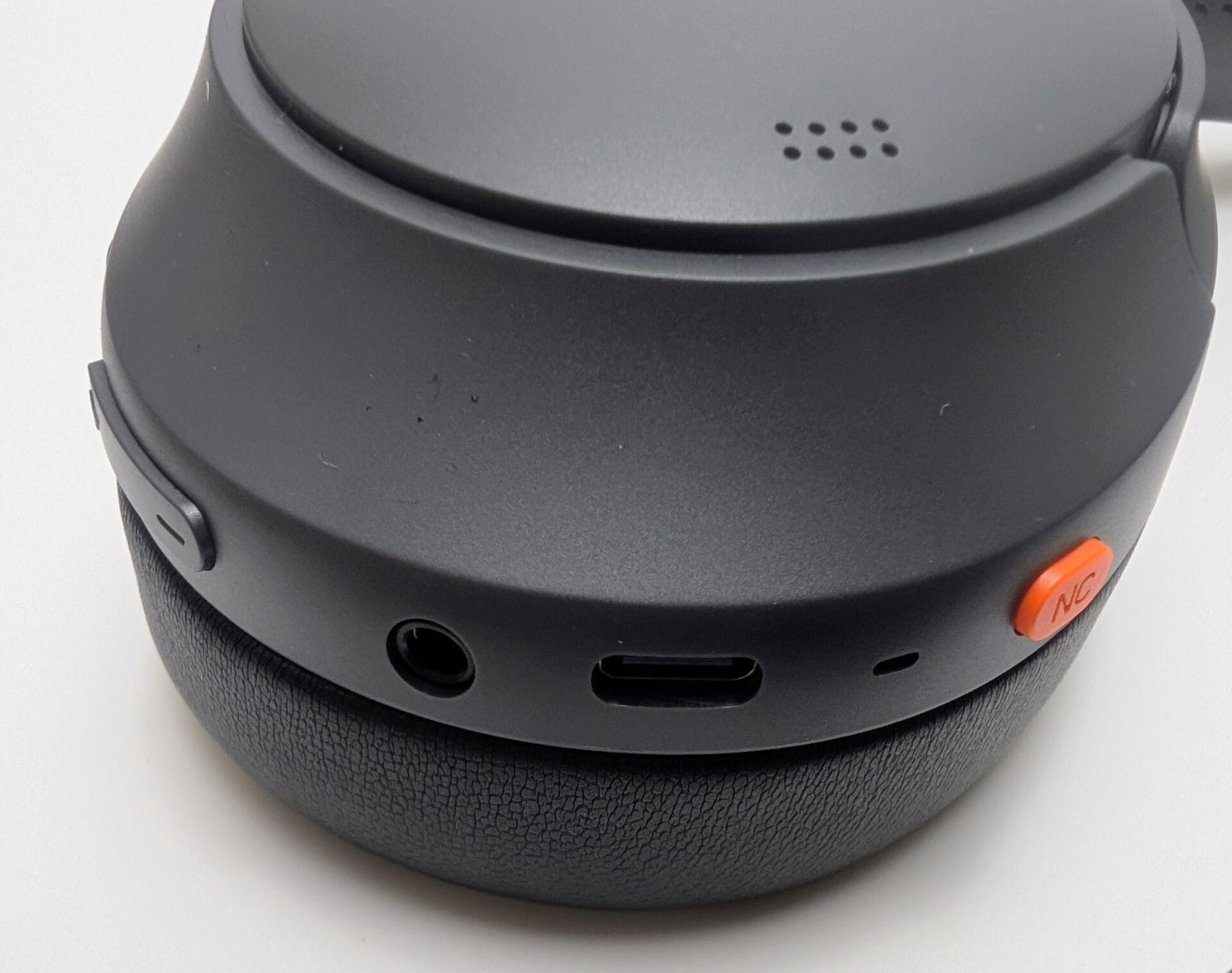 The image shows a close-up view of the right ear cup of the TOZO HT3 Adaptive Hybrid Active Noise Cancelling Headphones, focusing on the bottom edge and controls. The matte black ear cup has a rounded, ergonomic design with a textured protein leather ear pad at the base. On the lower side of the housing, visible from an angled perspective, are:A small 3.5mm AUX audio input jack (for wired listening).A USB-C charging port positioned centrally. A tiny microphone hole or vent nearby. A prominent bright orange button labeled "NC" (for toggling Active Noise Cancellation modes, Transparency mode, and holding for Low Latency mode). Above these, on the outer surface of the ear cup lid, there is a cluster of small perforations (likely for additional microphones or sound vents). The overall finish is smooth matte black with subtle branding absence in this cropped view, highlighting the practical physical controls and connectivity ports in a clean, user-friendly layout. This detailed shot emphasizes the headphone's hybrid wired/wireless capabilities and easy ANC switching