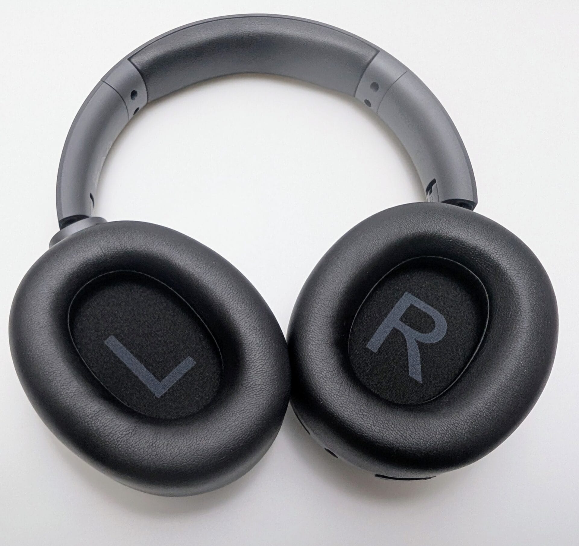 The image shows a close-up front view of the TOZO HT3 Adaptive Hybrid Active Noise Cancelling Headphones laid flat on a light surface, facing directly toward the camera. The matte black over-ear headphones feature a smoothly curved, padded headband in dark gray with subtle adjustment sliders and small ventilation holes near the hinges. The large, round ear cups are covered in textured black protein leather with plush, cushioned padding. The interior of the left ear cup displays a faint gray "L" marking near the center, while the right ear cup shows a matching "R" marking, clearly indicating left and right sides. The "TOZO" branding is absent from the ear pads in this view, emphasizing the minimalist design with subtle perforations along the outer edges of the ear cups for microphones or vents. The overall appearance is sleek, modern, and comfortable-looking, with a premium matte finish throughout.