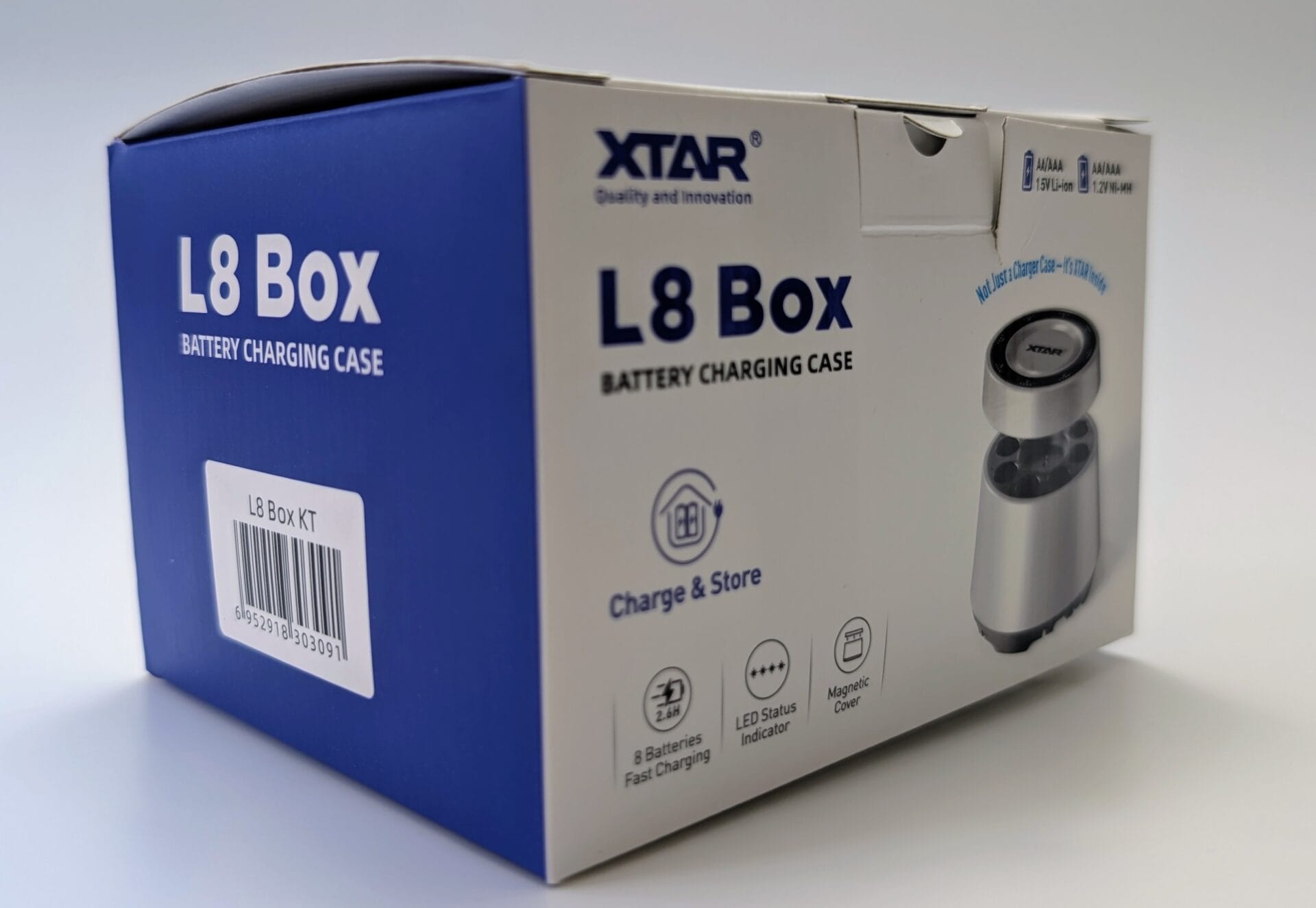 XTAR L8 Box Charger A close-up product photograph of the XTAR L8 Box Battery Charging Case packaging. The blue and white cardboard box prominently displays 'L8 Box BATTERY CHARGING CASE' on both the blue side and white front panels. The front features an image of the sleek silver cylindrical charging case, along with icons and text highlighting features such as 'Charge & Store', '8 Batteries Fast Charging', '2.8H', 'LED Status Indicator', and 'Magnetic Cover'. A barcode label reading 'L8 Box KT' is visible on the blue side. The packaging also shows compatibility icons for AA/AAA 1.5V Li-ion and 1.2V Ni-MH batteries.