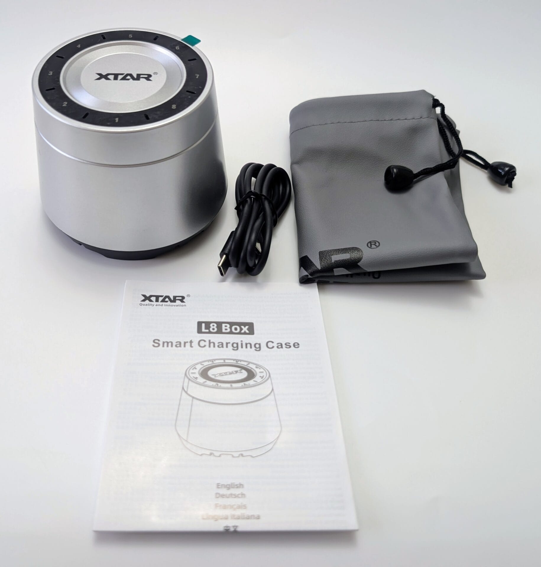 XTAR L8 Box Charger An overhead shot of the XTAR L8 Box Smart Charging Case kit on a white background. The set includes a cylindrical silver charging unit with eight numbered battery slots on top, a black USB-C charging cable, a grey drawstring storage pouch with the XTAR logo, and a printed user manual.