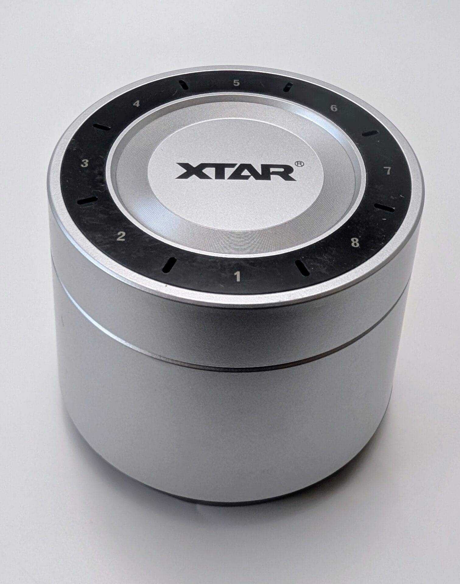 XTAR L8 Box Charger A close-up, high-angle shot of the XTAR L8 Box Smart Charging Case. The device is a sleek, silver cylindrical unit with the "XTAR" logo centered on the top. A black circular ring around the top features numbered slots from 1 to 8, each with a small indicator light, designed for charging multiple batteries simultaneously.
