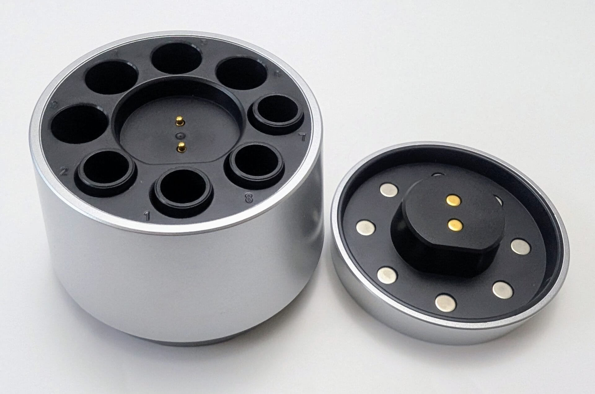 XTAR L8 Box Charger An overhead view of the XTAR L8 Box charger with its magnetic lid removed and placed to the right. The silver cylindrical base reveals eight circular battery charging bays arranged in a ring. The center of the base and the underside of the lid both feature gold-plated pogo pin connectors and magnetic alignment points for secure closing.