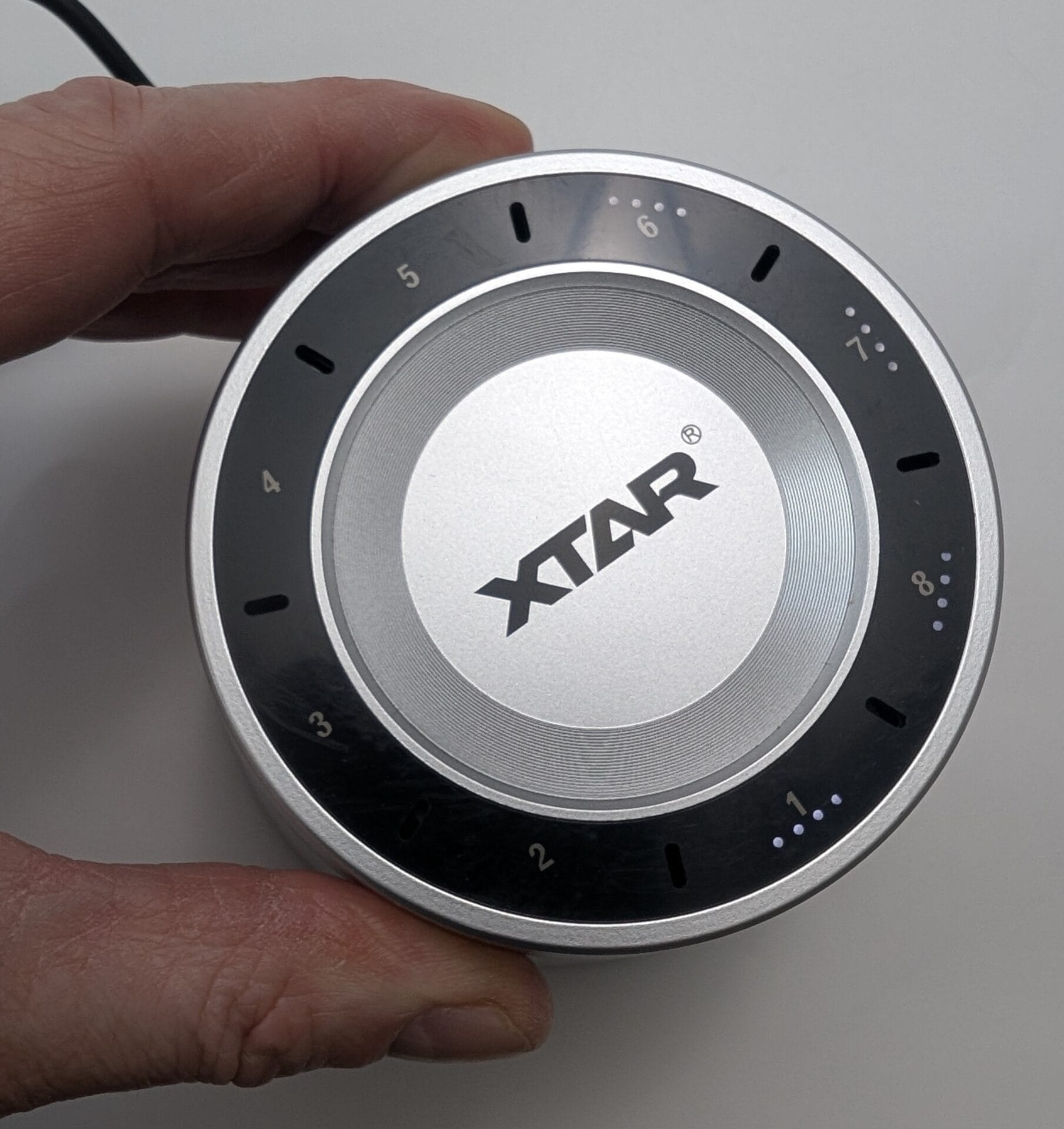 XTAR L8 Box Charger A top-down close-up of the XTAR L8 Box charger being held by a hand. The black circular interface is illuminated, showing white LED dot indicators next to the numbers. Slot 1 shows four dots, while slots 6, 7, and 8 show varying levels of charge, demonstrating the real-time battery status monitoring feature.