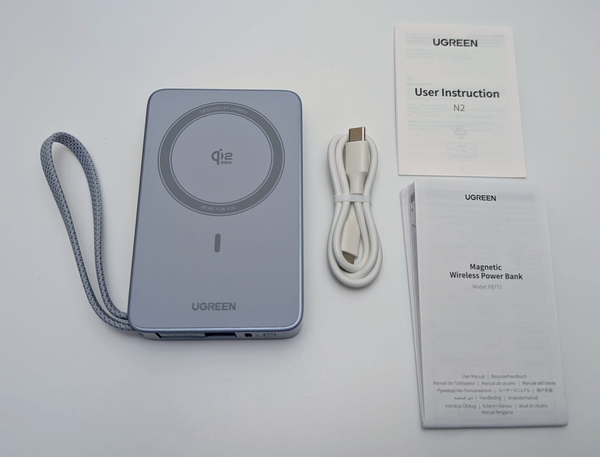 An unboxed UGREEN MagFlow 10000mAh Magnetic Wireless Power Bank (Model PBT73) with a 25W Qi2-certified wireless charging pad. The image shows the sleek gray power bank with a circular charging area, a built-in braided strap, and a USB-C port. Included are a white USB-C cable and user instruction manuals in multiple languages.