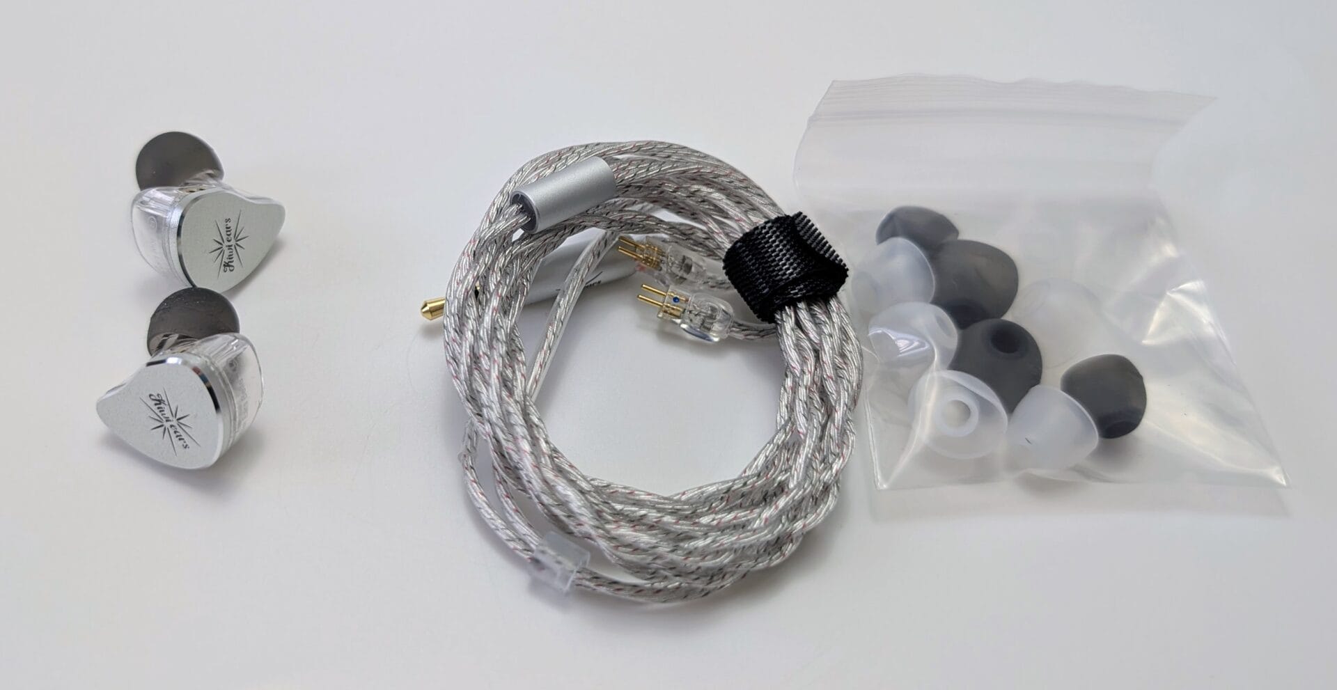 Contents of the Kiwi Ears Belle in-ear monitors (IEMs) laid out on a white surface. On the left are the two silver metallic earpieces with the Kiwi Ears starburst logo. In the center is the coiled silver braided cable with a 3.5mm gold-plated plug and two-pin connectors. On the right is a clear plastic bag containing multiple pairs of silicone and foam eartips in black, gray, and translucent colors.
