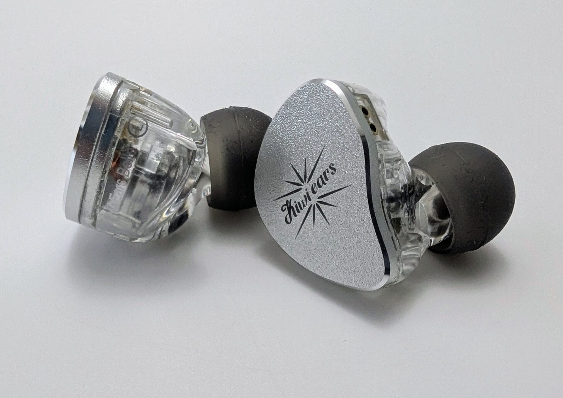 Pair of Kiwi Ears Belle in-ear monitors (IEMs) resting on a white surface. The left earpiece is shown from the side, revealing its transparent acrylic shell and internal components, while the right earpiece displays the brushed silver faceplate with the engraved Kiwi Ears starburst logo. Both have black silicone eartips attached.